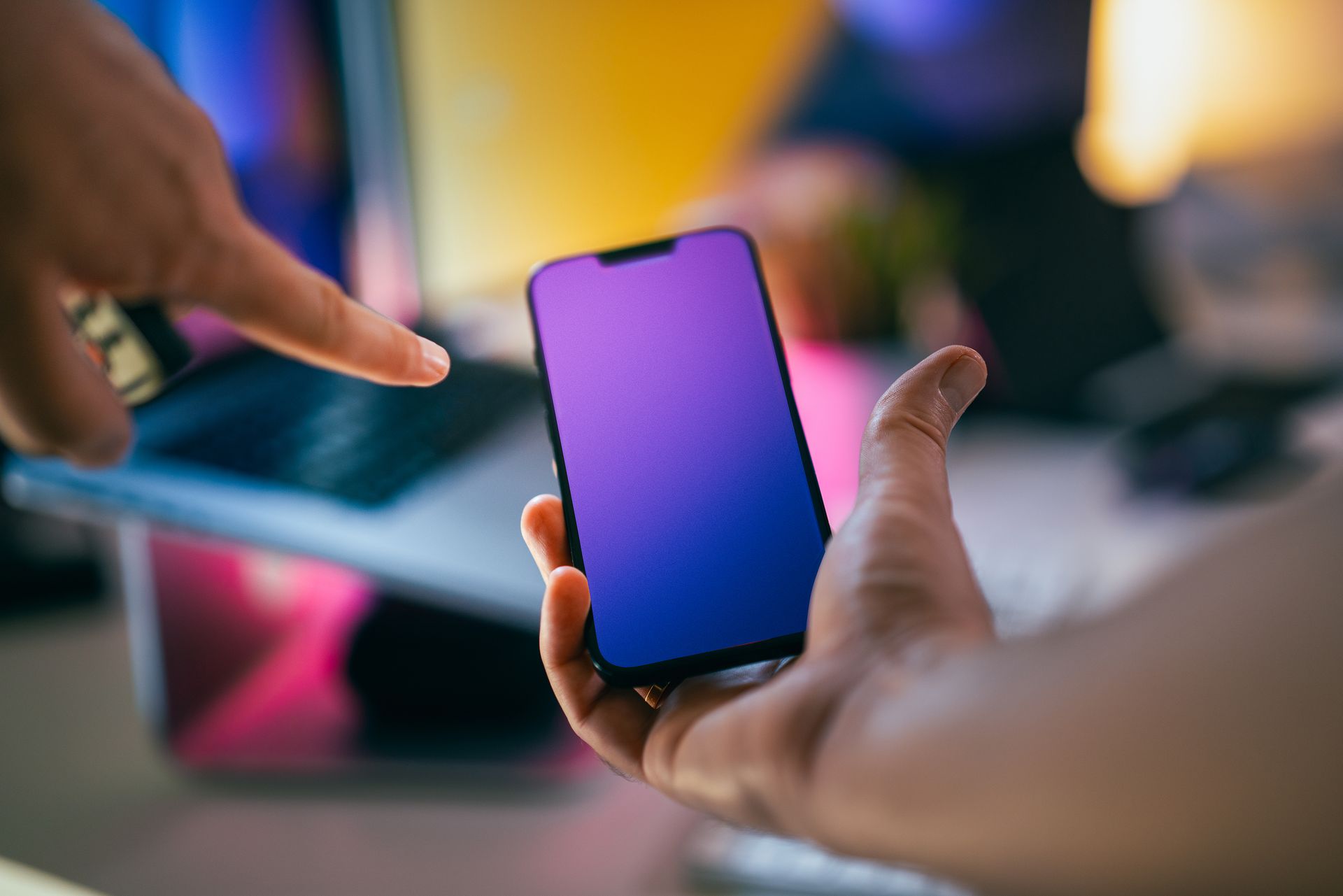 Hands pointing at a smartphone with a gradient screen in an office setting.