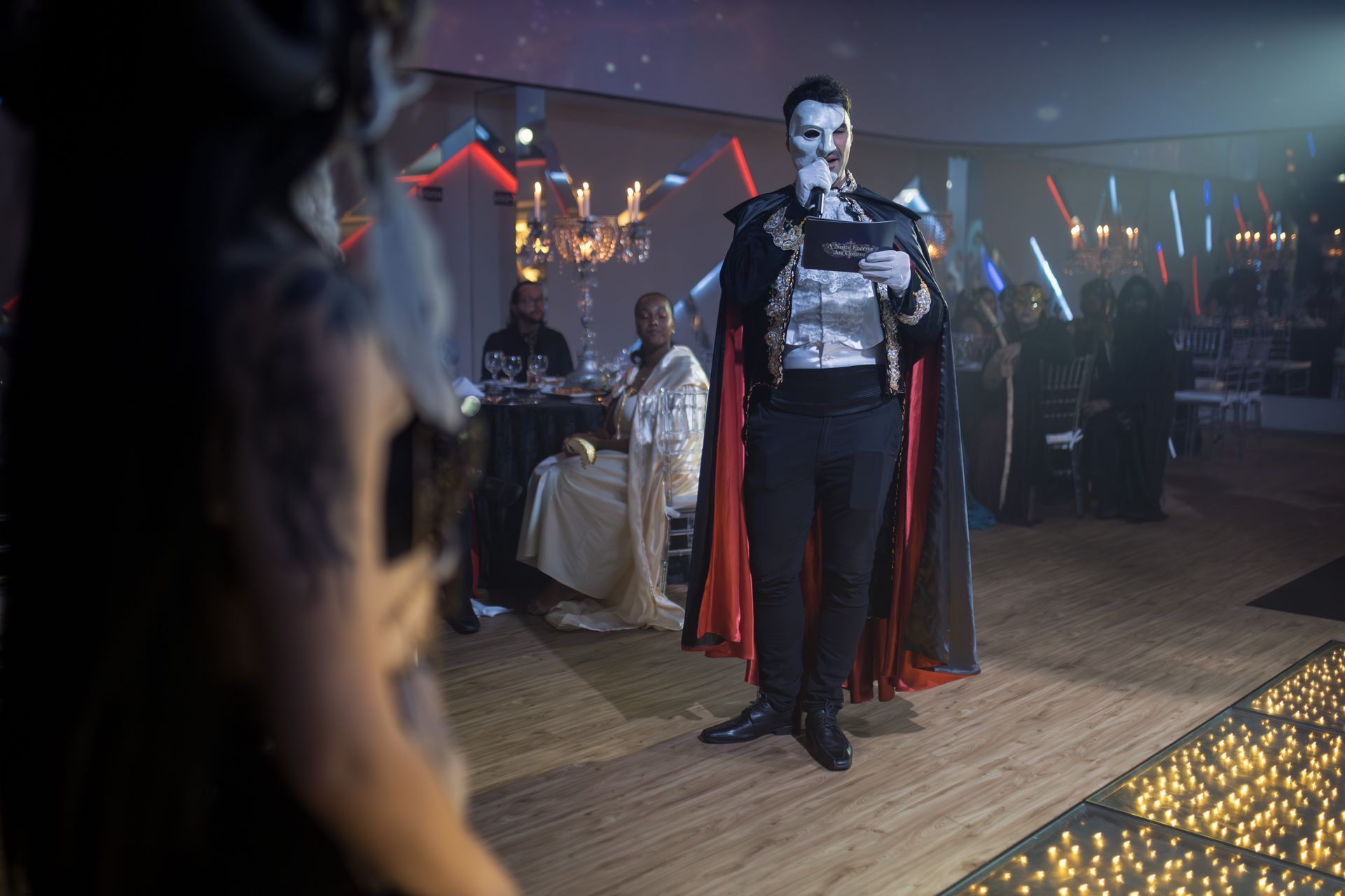 A masked host in formal attire speaks at a glamorous costume party event.