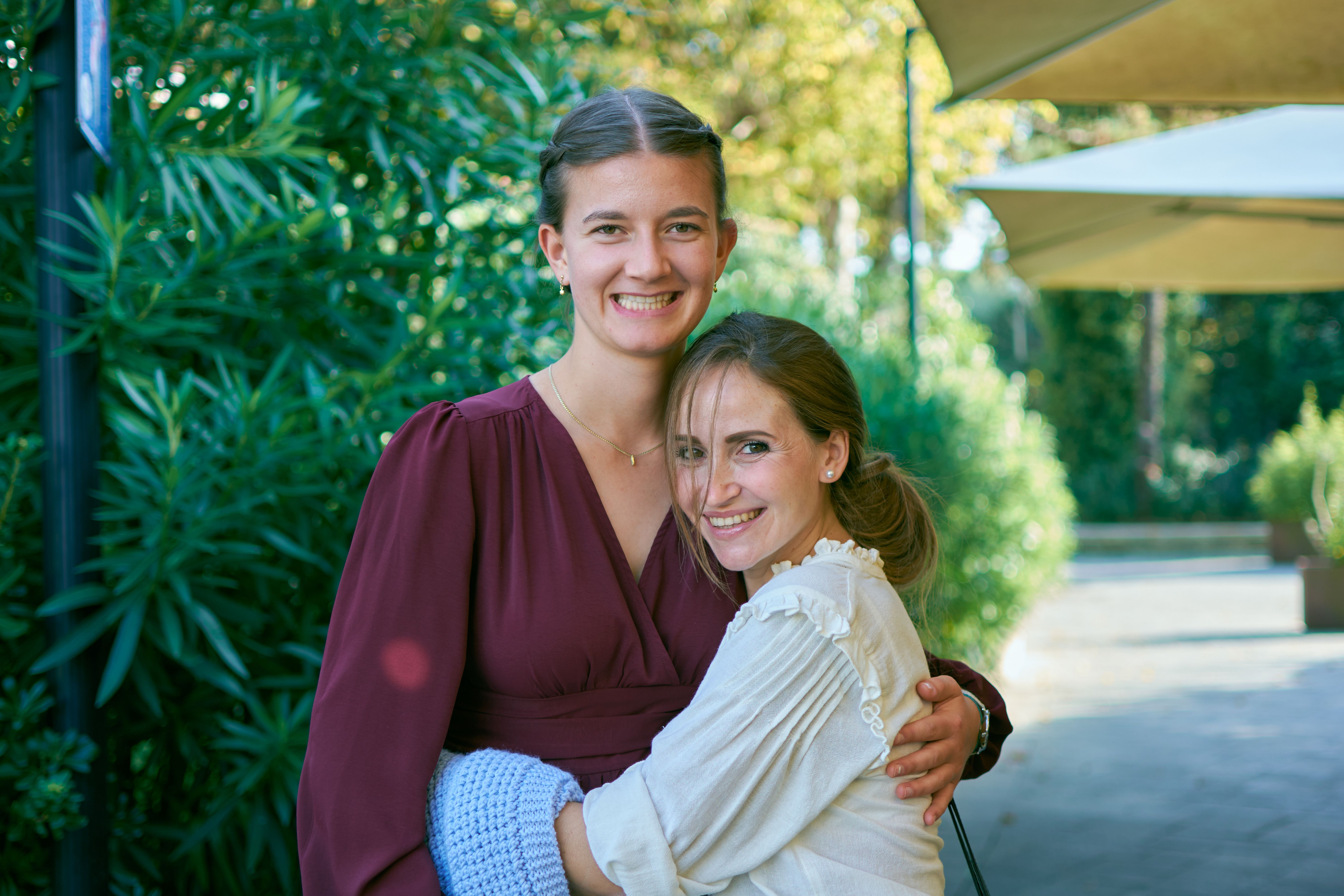 Two women smiling and embracing outdoors in a sunny setting.