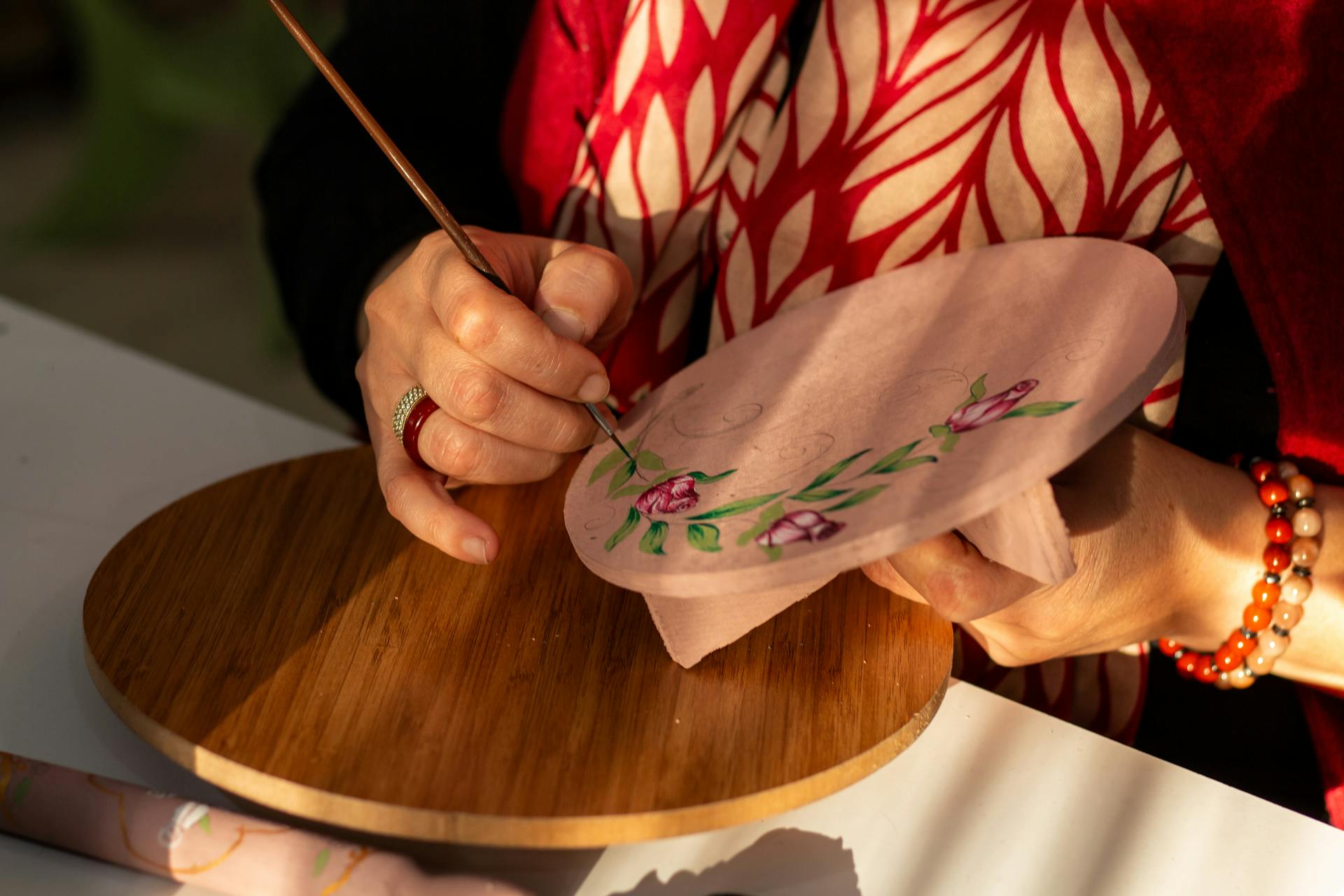 Close-up of an artist painting a floral design on a round canvas board with sunlight.