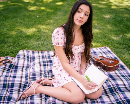 A young woman sitting on a picnic blanket and writing in a notebook outdoors.