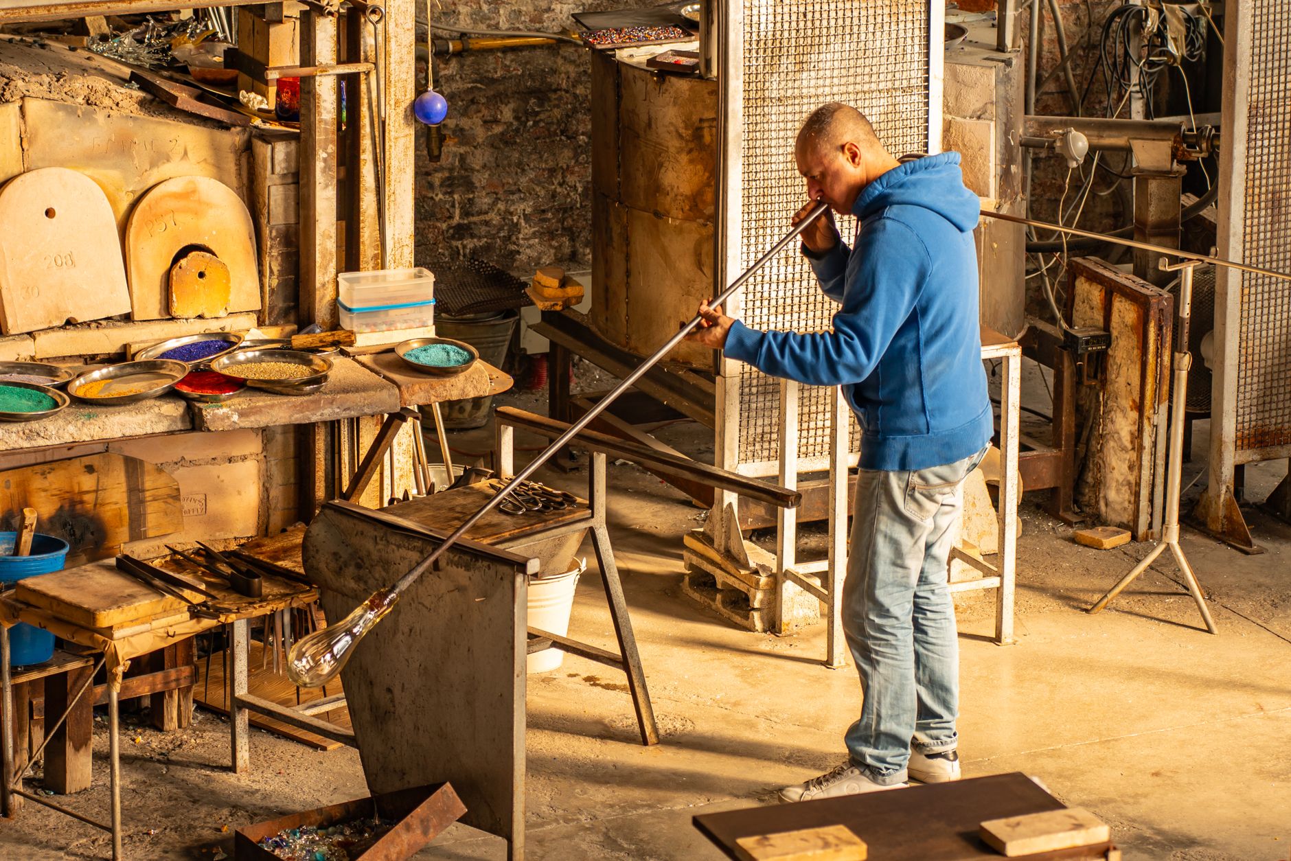A glassmaker gathering molten glass on a blowing iron in a small workshop, trays of coloured frit on the bench