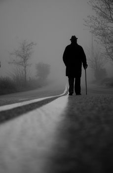 A solitary elderly man with a cane walks on an empty, fog-covered road, creating a moody silhouette.