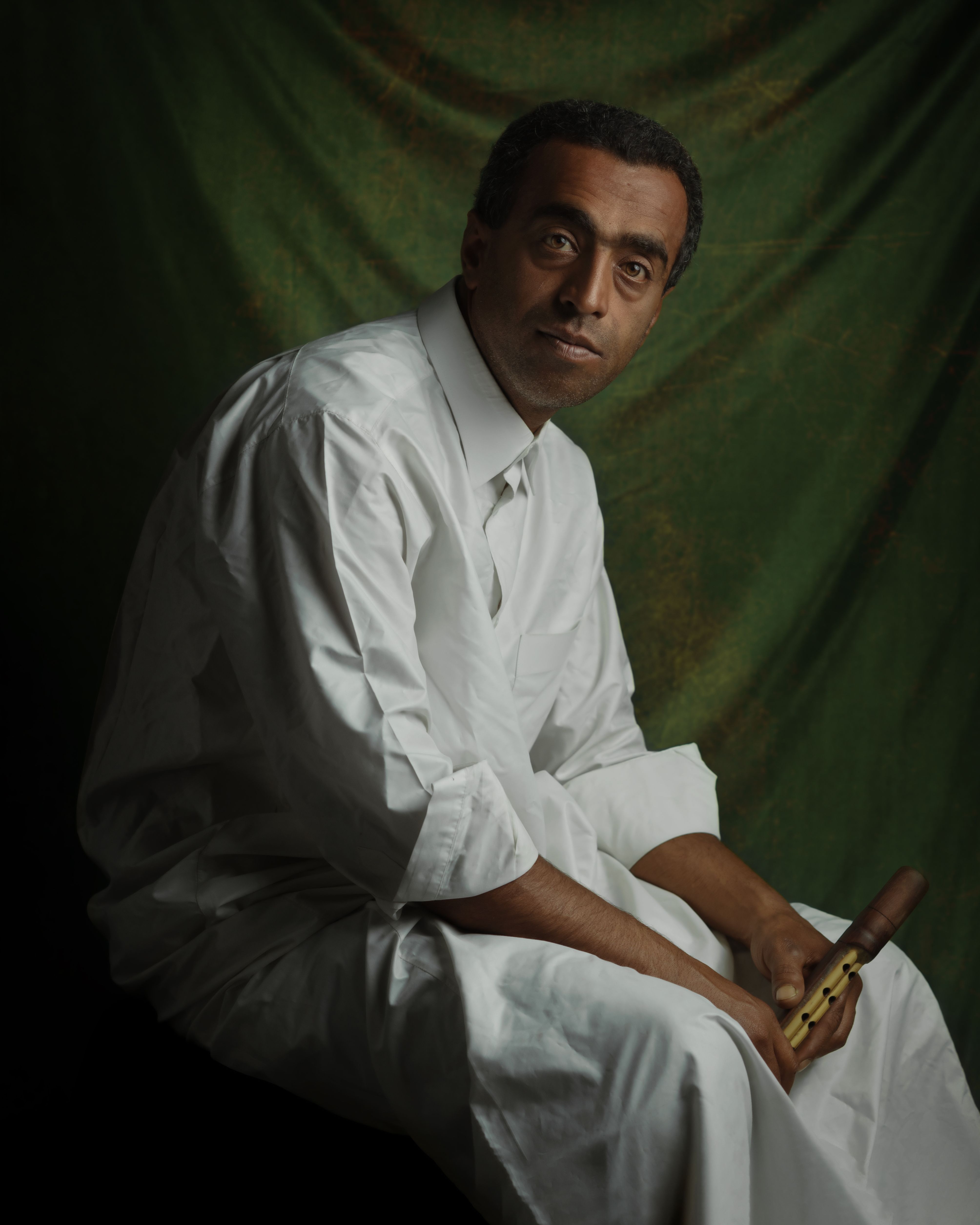 A thoughtful man in a white robe holding a traditional flute against a green backdrop.