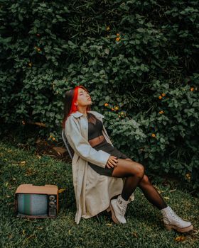 Fashionable woman in trench coat posing with retro TV in a lush garden setting, exuding a nostalgic vibe.