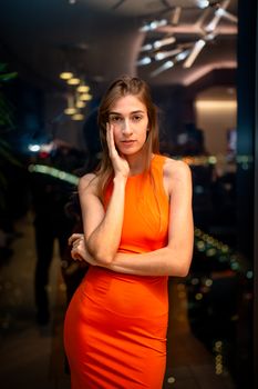 Stylish woman in an orange dress posing indoors with a city night view.