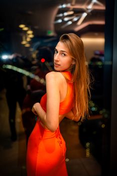 Stylish woman in a red dress posing indoors against a city backdrop at night.