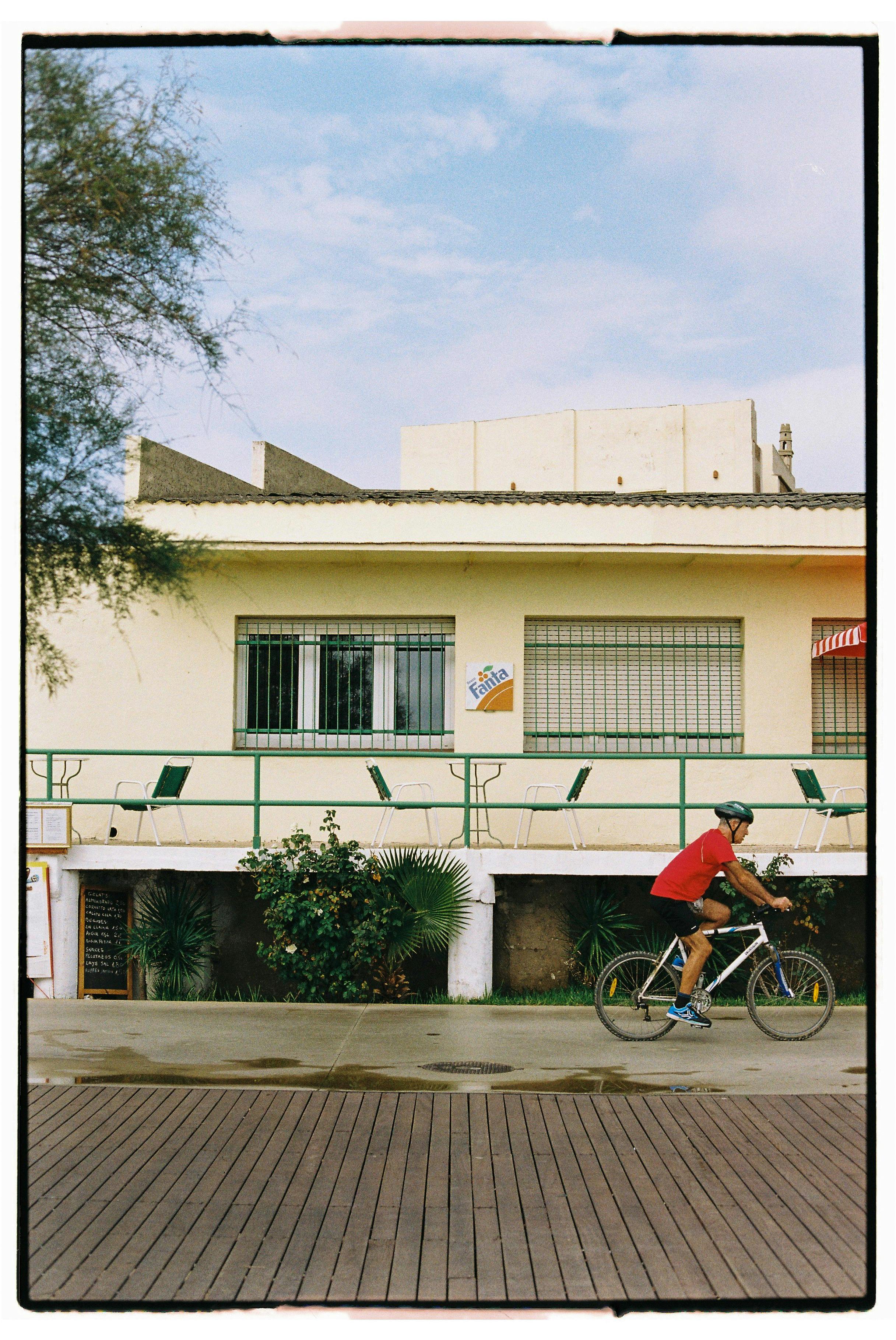 A cyclist in a red shirt rides past a yellow building on a sunny day.