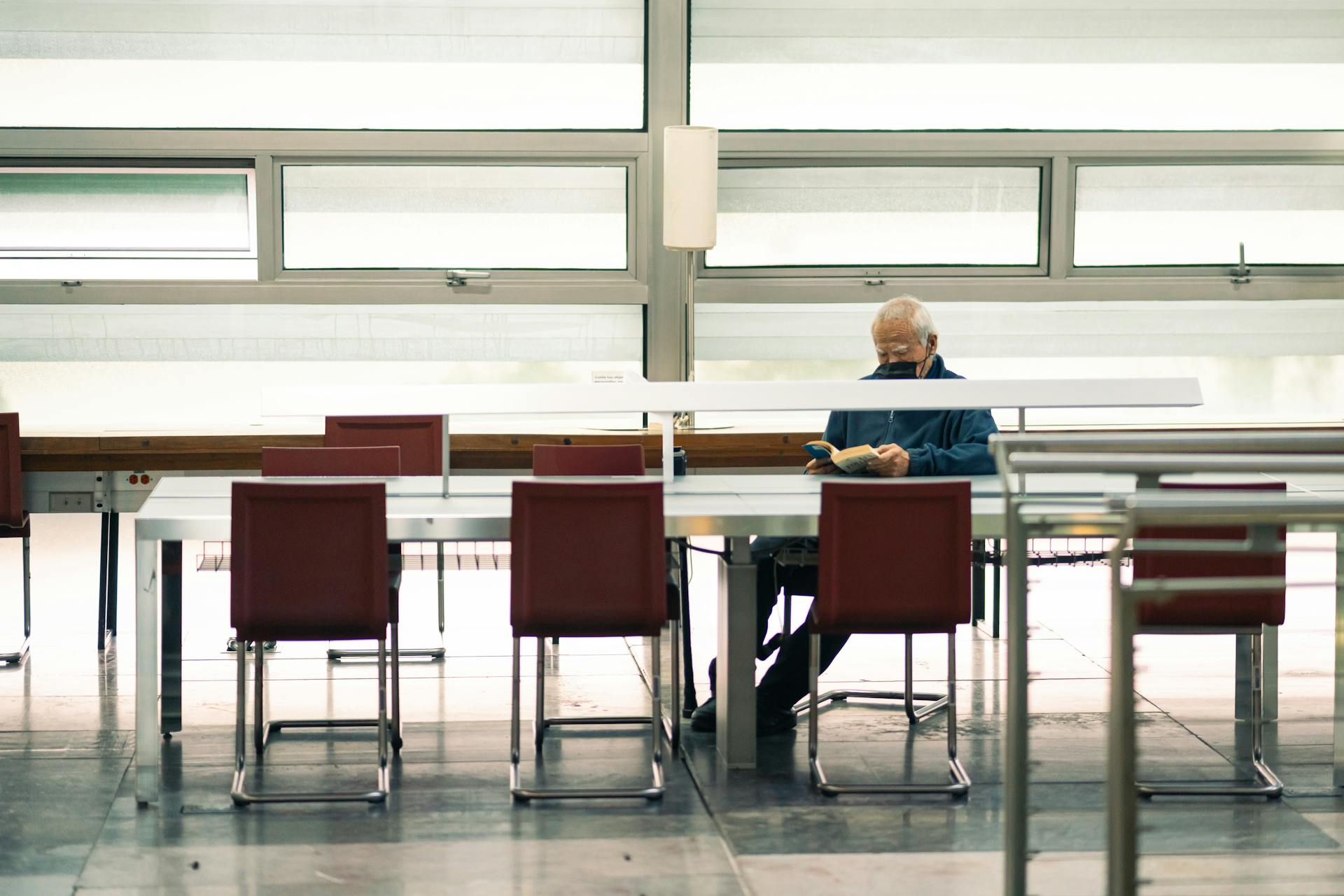 Elderly man reading book in quiet modern library with natural light.