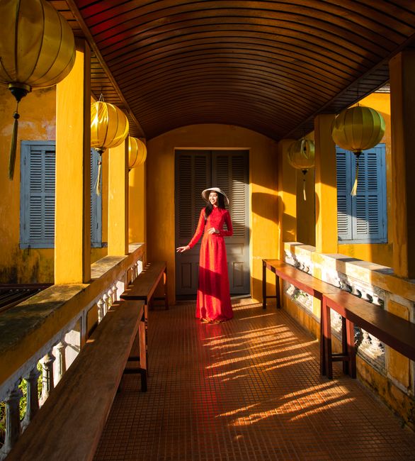 Woman in red dress and hat standing in a sunlit corridor of Hoi An, Vietnam.