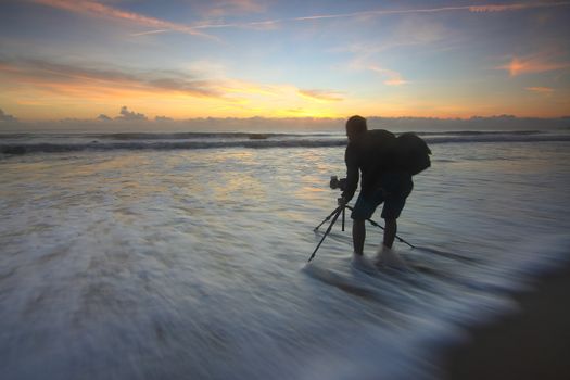 A photographer takes a long exposure shot at sunrise on a beach with waves crashing.