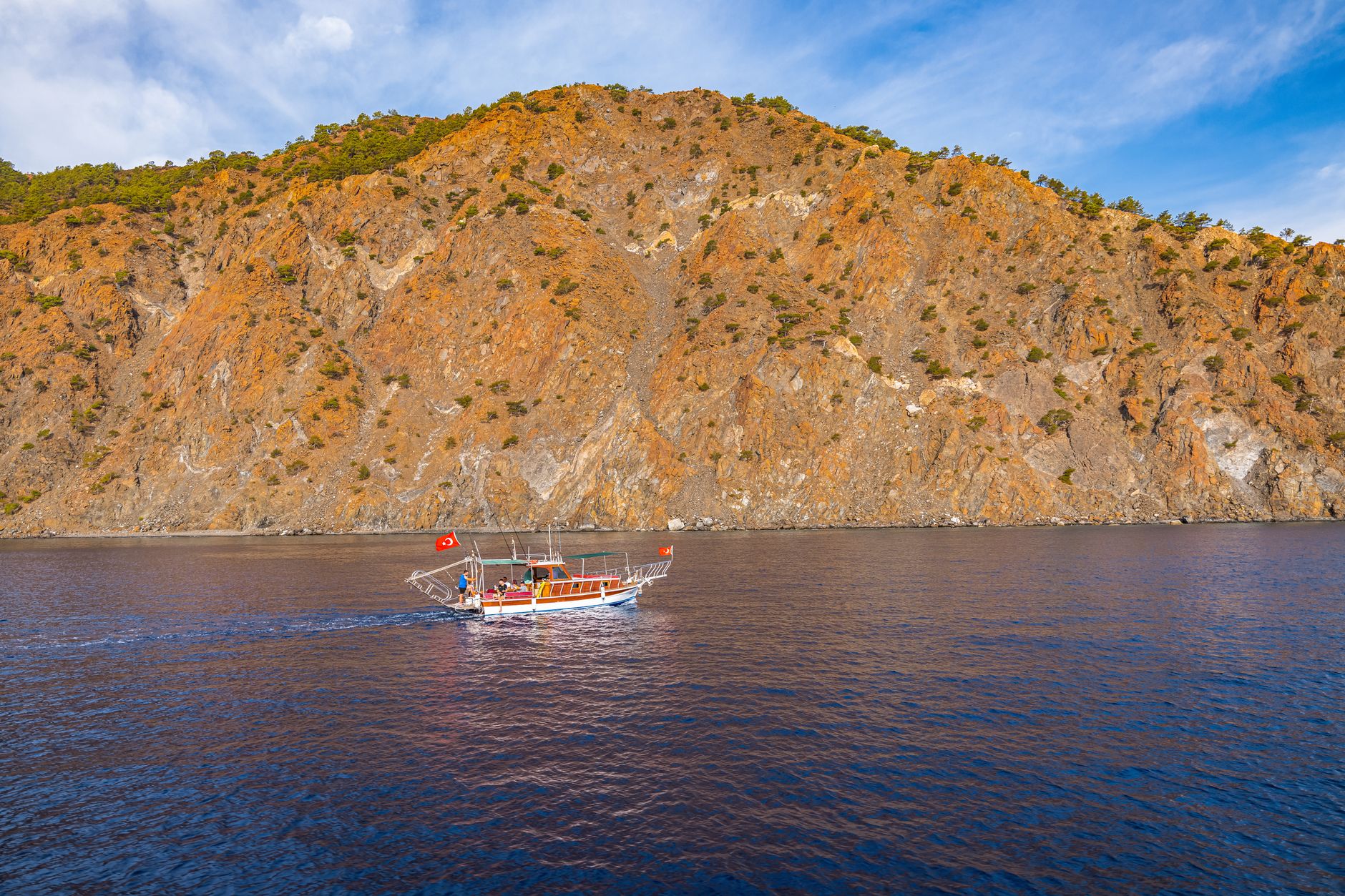 A boat with a Turkish flag cruises along a rocky coastline under a clear sky.