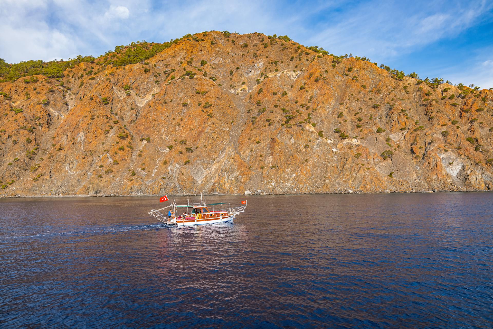 A boat with a Turkish flag cruises along a rocky coastline under a clear sky.