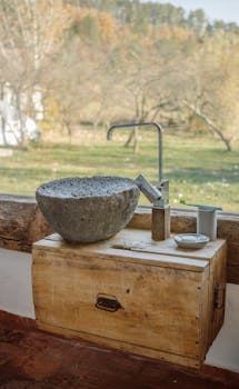 A rustic stone sink on a wooden pedestal, overlooking a serene outdoor landscape through a window.