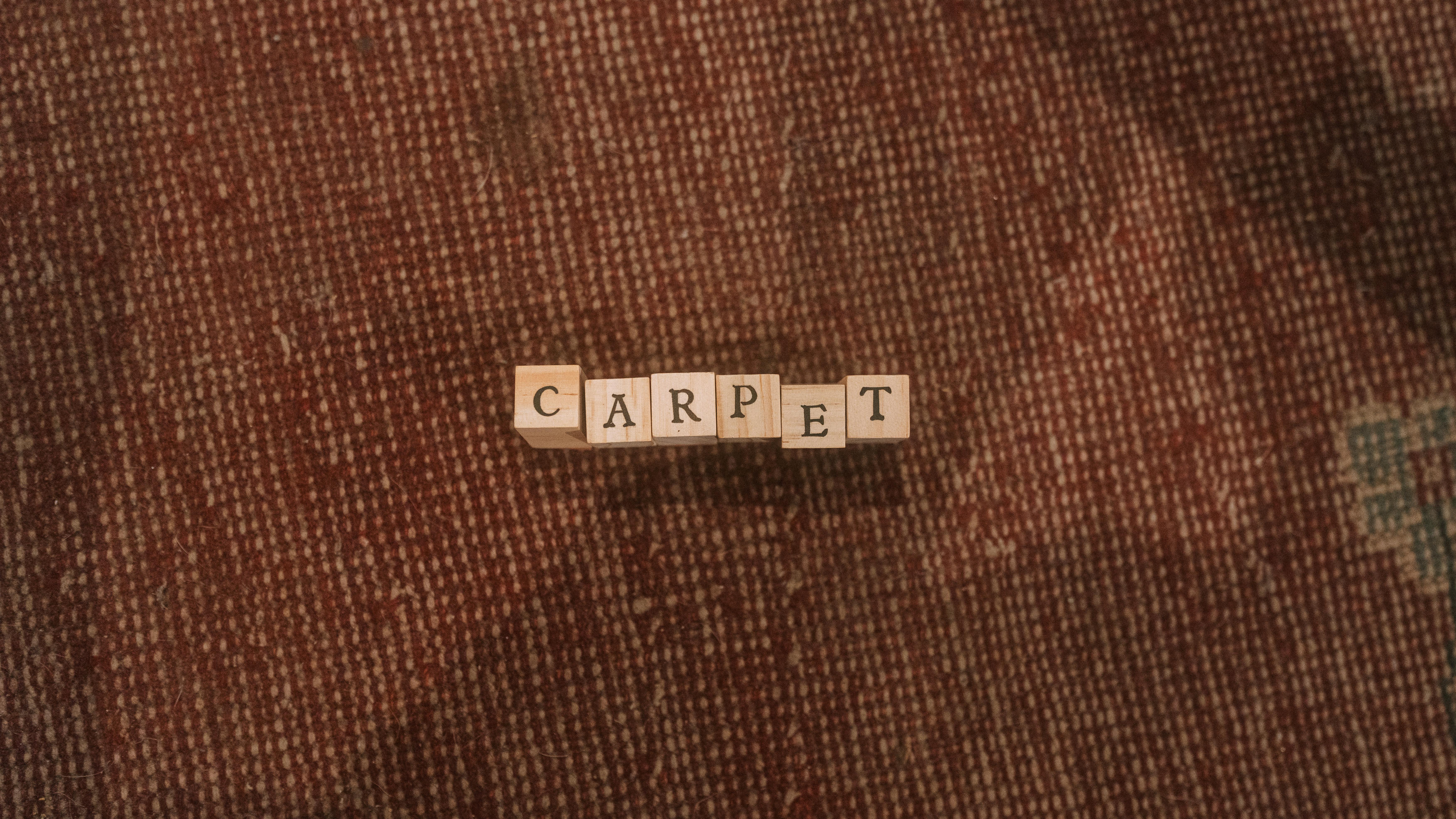 Free Wooden letter tiles spell 'carpet' on a textured rug surface. Simple and warm composition. Stock Photo