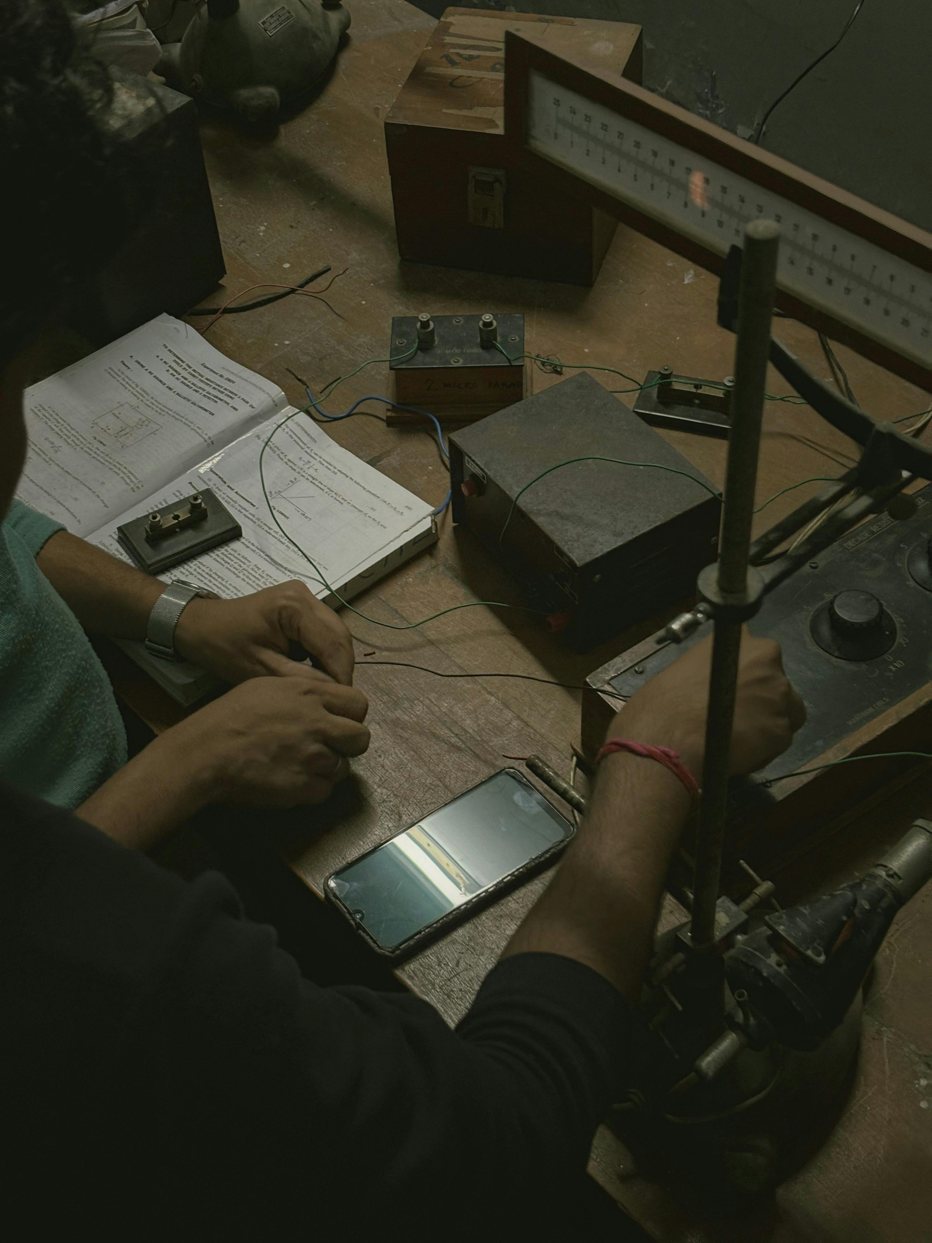 Hands working on electronics project with equipment and notes in a dimly lit workshop.