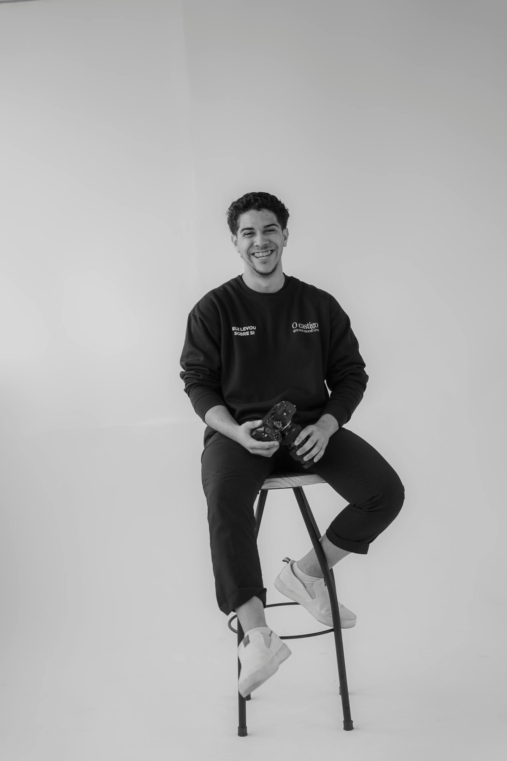 Gratis Ritratto in bianco e nero di un uomo sorridente seduto su uno sgabello con in mano una macchina fotografica. Foto a disposizione