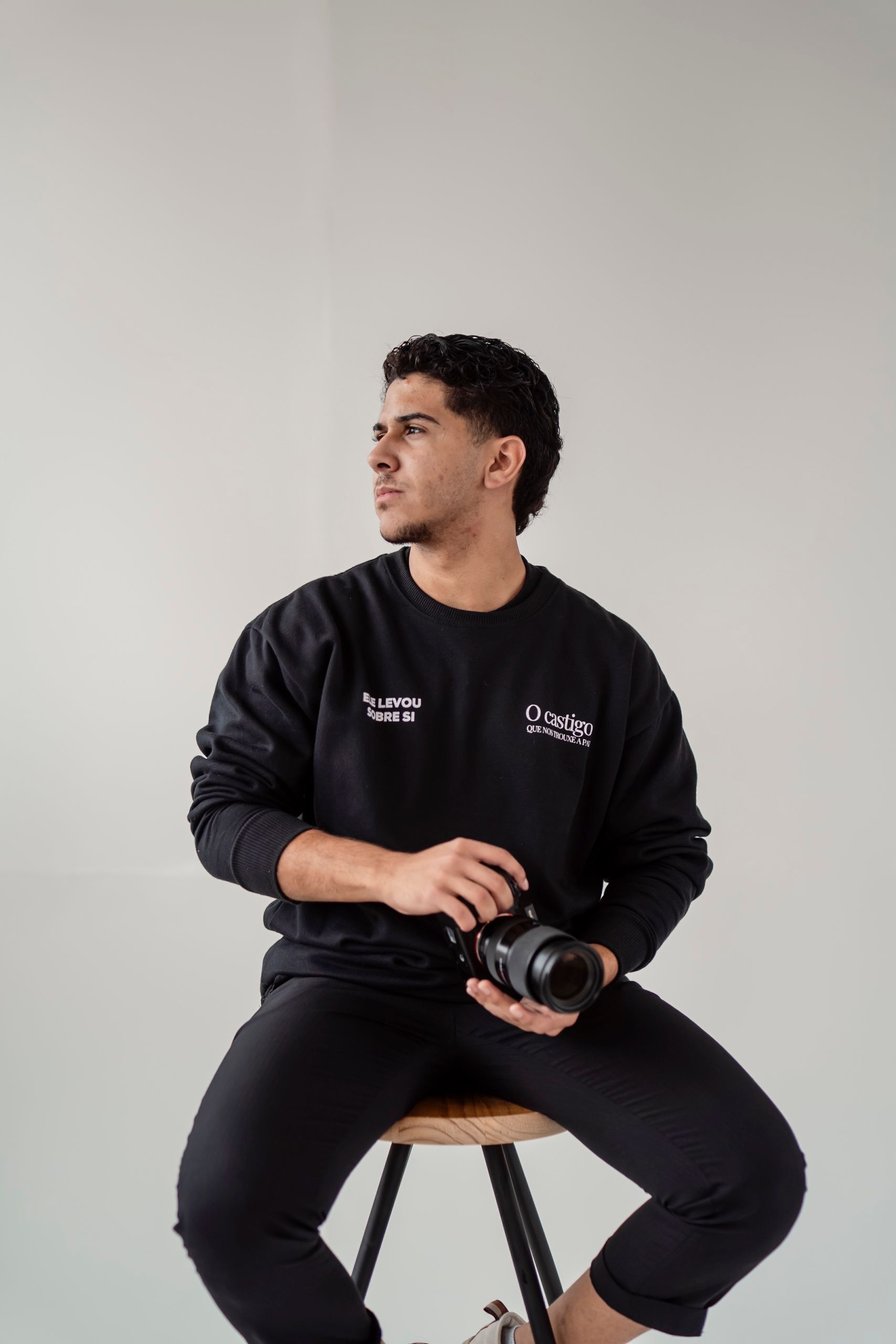 Portrait of a young man holding a camera, sitting on a stool in a studio setting.
