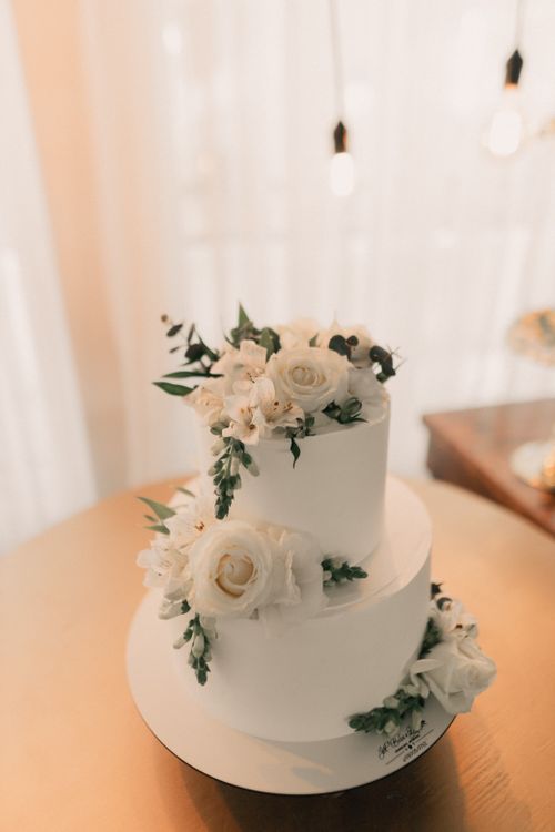 Free Beautiful two-tier wedding cake adorned with white roses and greenery on a golden table. Stock Photo