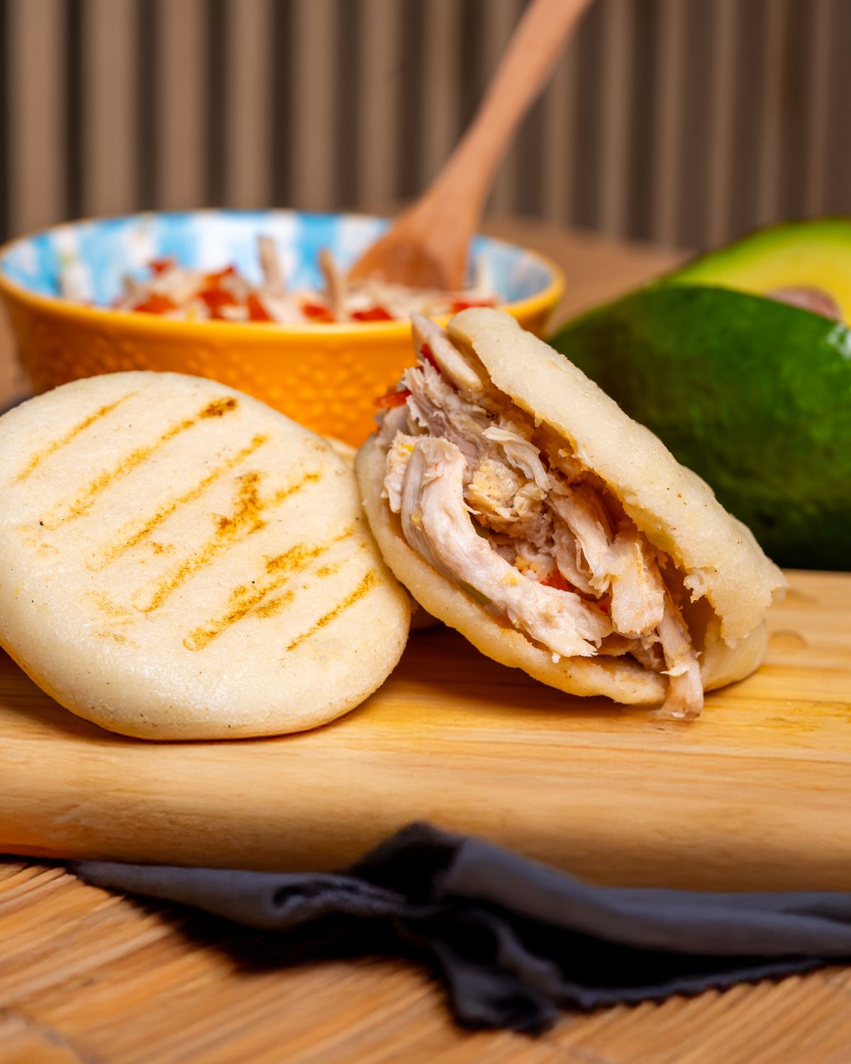 Grill-marked white corn arepa split and stuffed with shredded chicken beside an avocado
