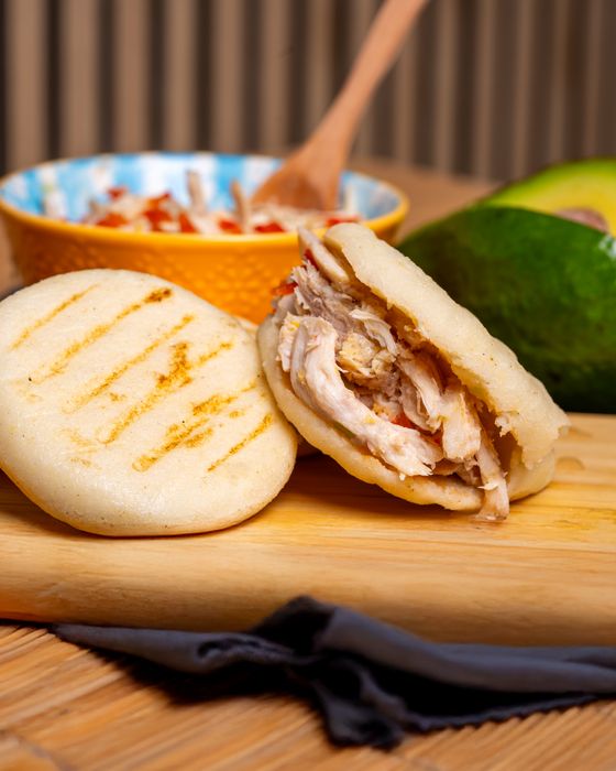 Grill-marked white corn arepa split and stuffed with shredded chicken beside an avocado