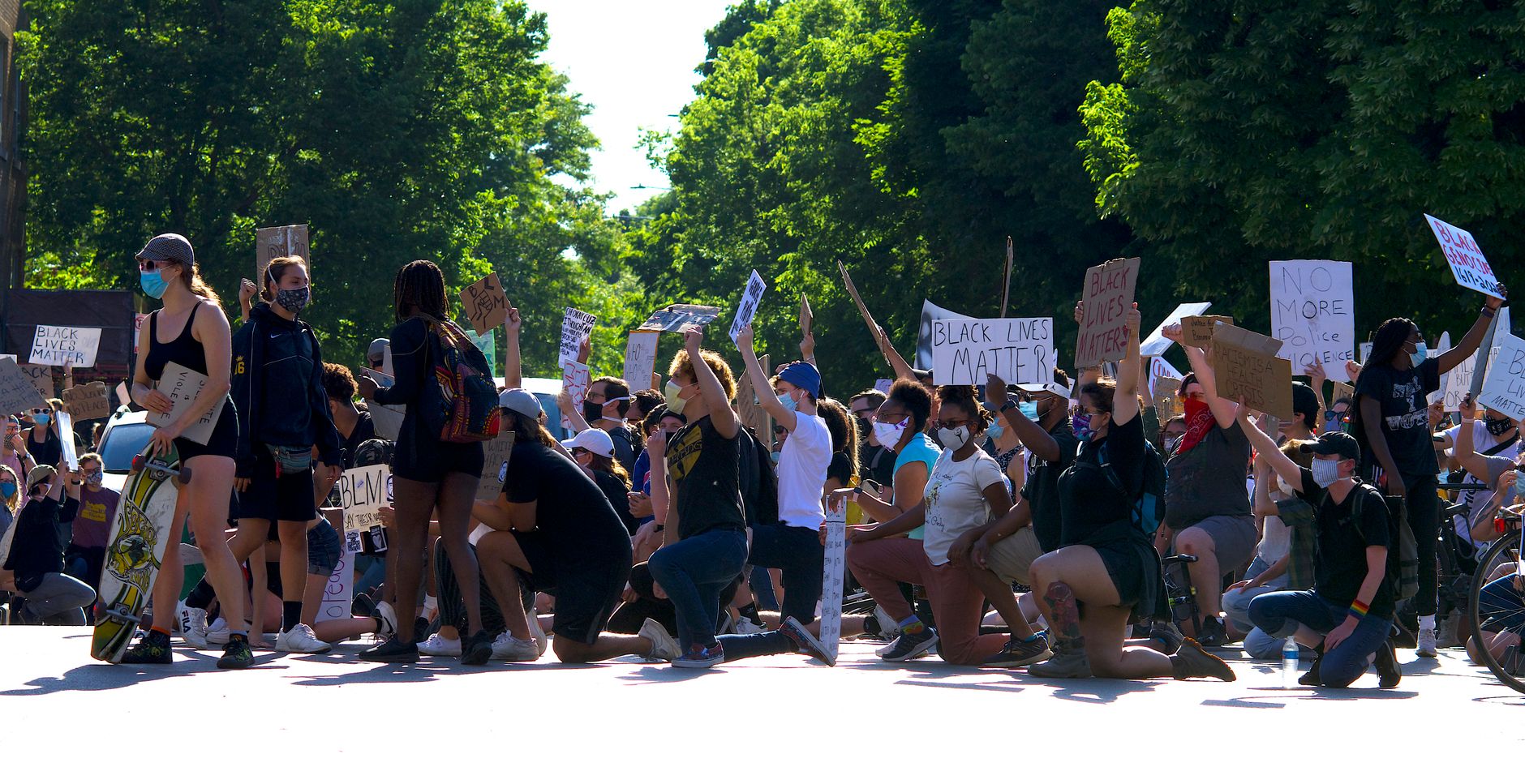 A group of diverse protesters kneel outdoors, holding signs for the Black Lives Matter movement.