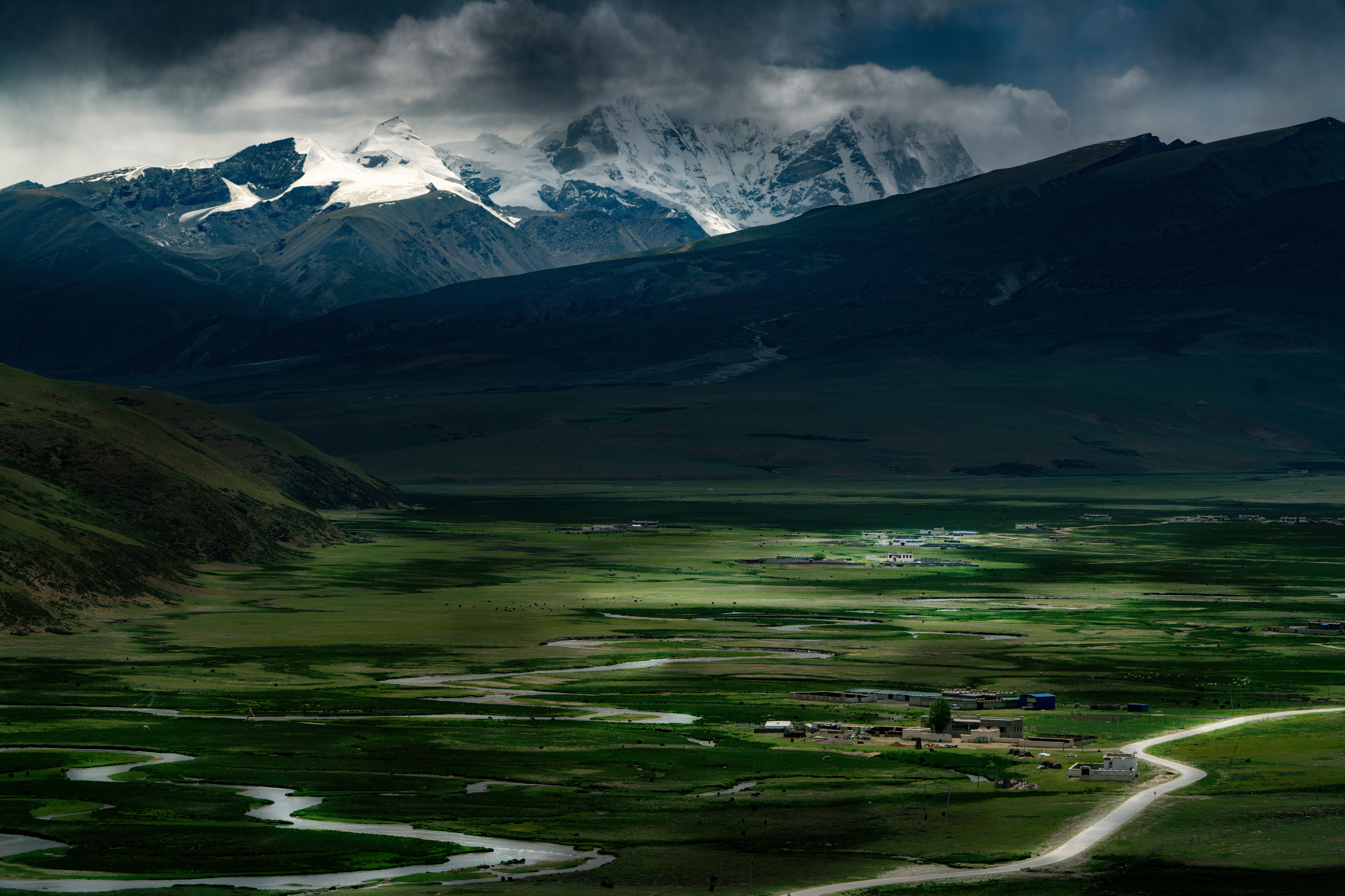 [ColoSach]-A breathtaking view of a green valley with snow-covered mountains under dramatic clouds.