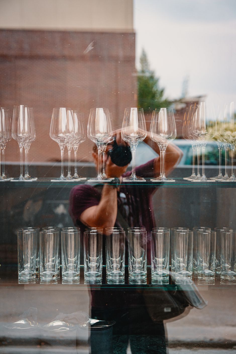 Reflection of a photographer capturing wine glasses and tumblers on a glass shelf.