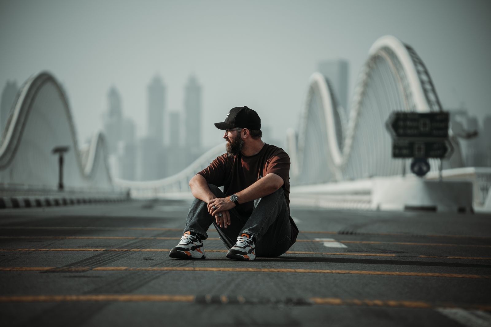 A person sits on the Meydan Bridge deck with its wave arches and the hazy Dubai skyline rising behind.