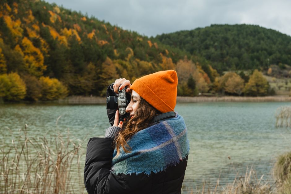 A woman in an orange beanie takes photos of a stunning autumn landscape by a lake.