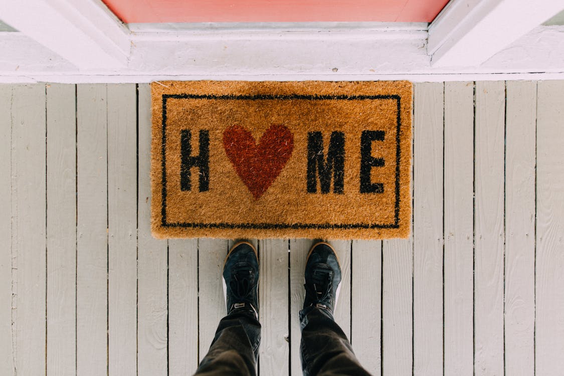 pov of person standing in front of a welcome mat with the word "Home" on it. The "O" in Home is replaced with a red heart. 