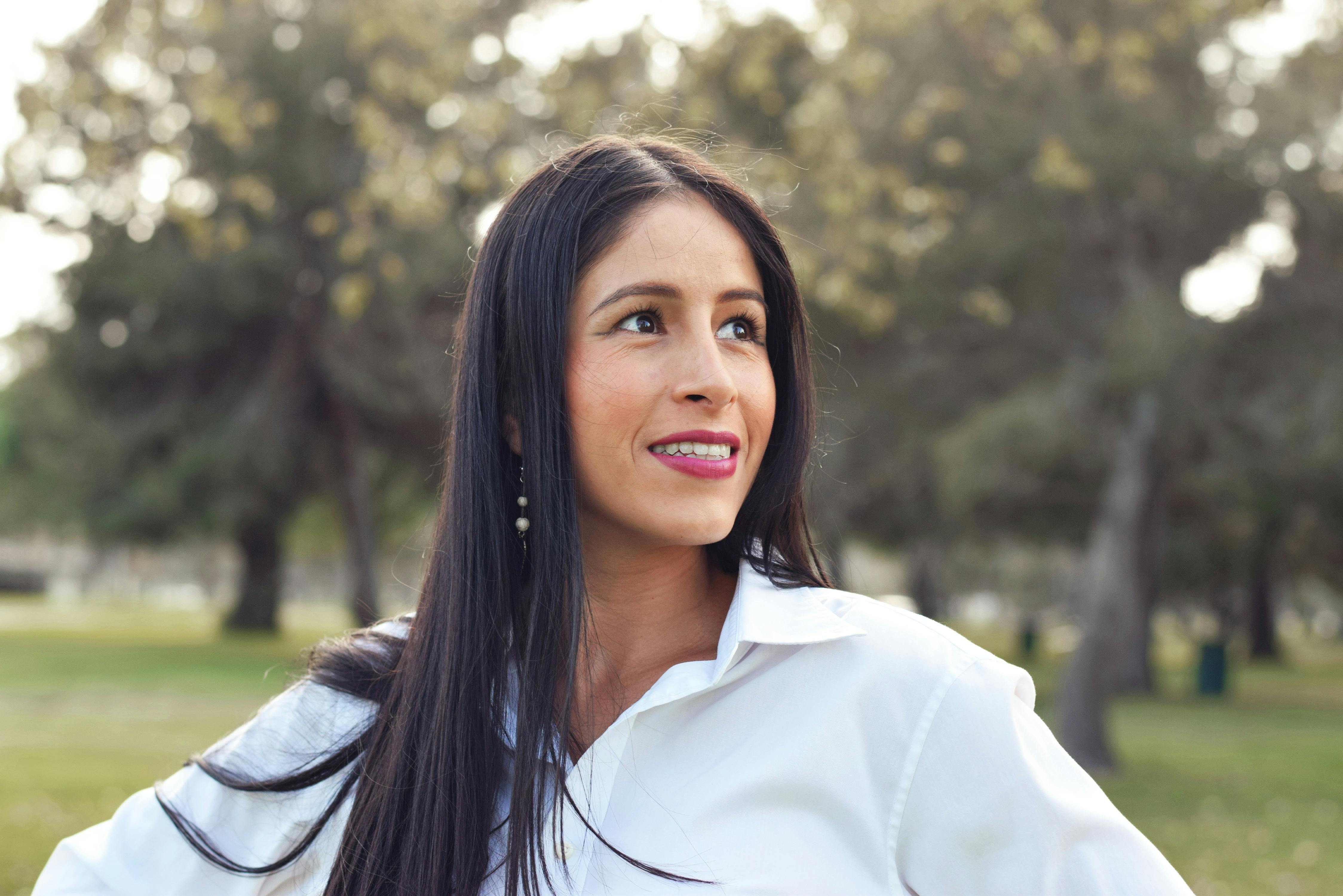 Ilmainen Pitkähiuksisen naisen muotokuva, joka poseeraa itsevarmasti auringon valaisemassa puistossa puiden ympäröimänä. Kuvapankkikuva