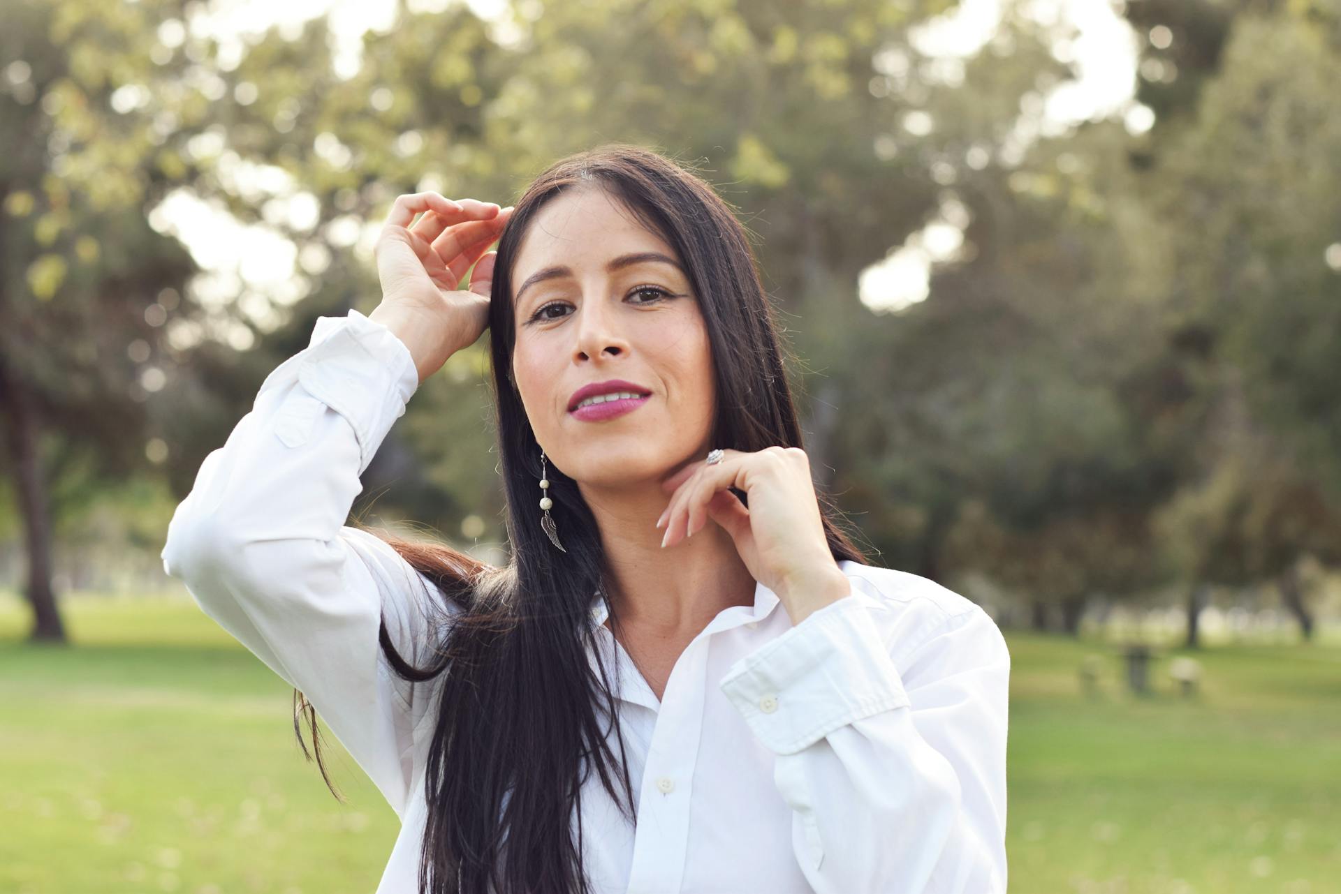 A woman in a white shirt poses gracefully outdoors in a Long Beach park with lush greenery.