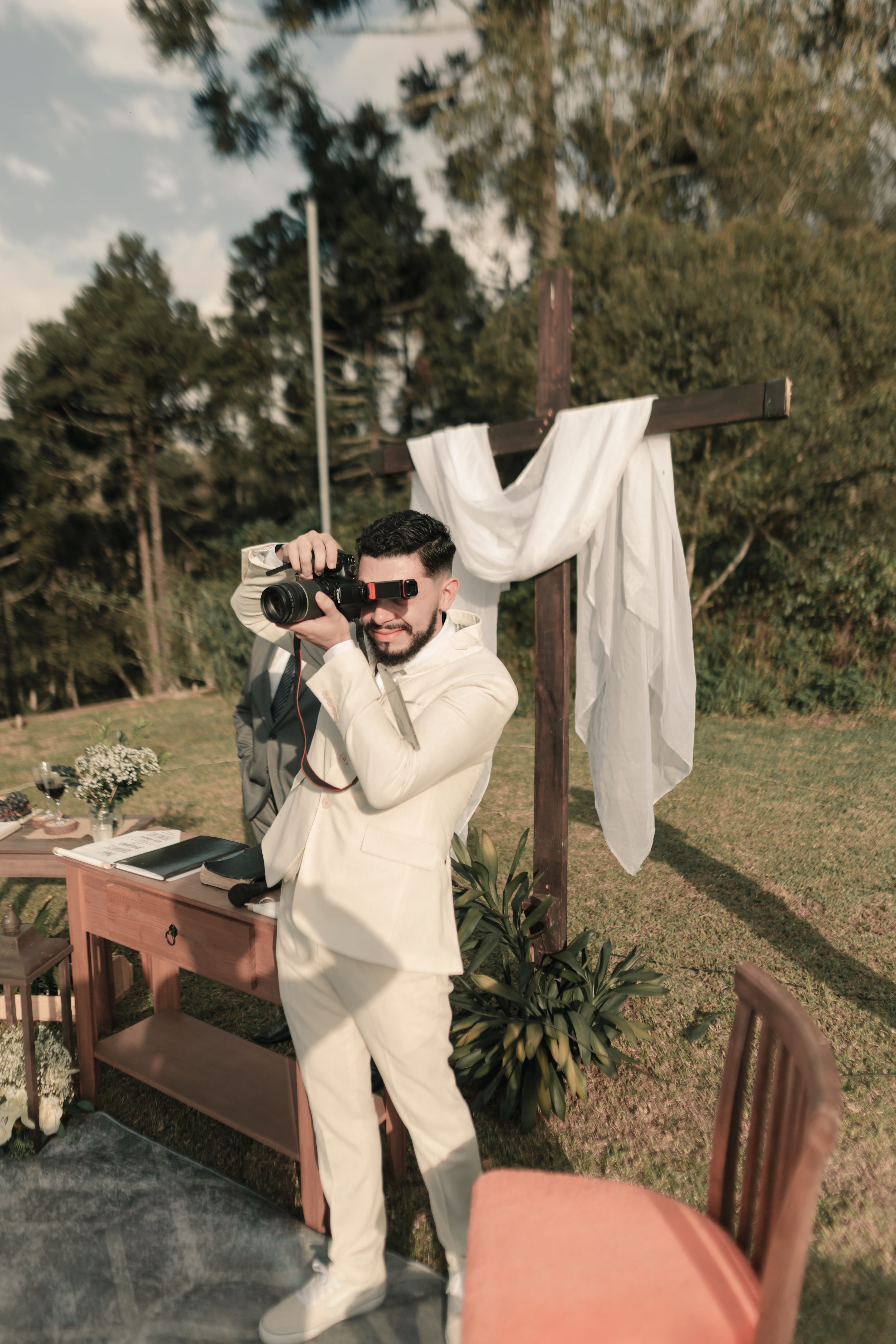 A photographer in a white suit takes pictures at an outdoor ceremony.