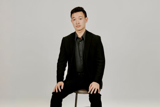 Formal portrait of a man seated on a stool, wearing a black suit against a white background.
