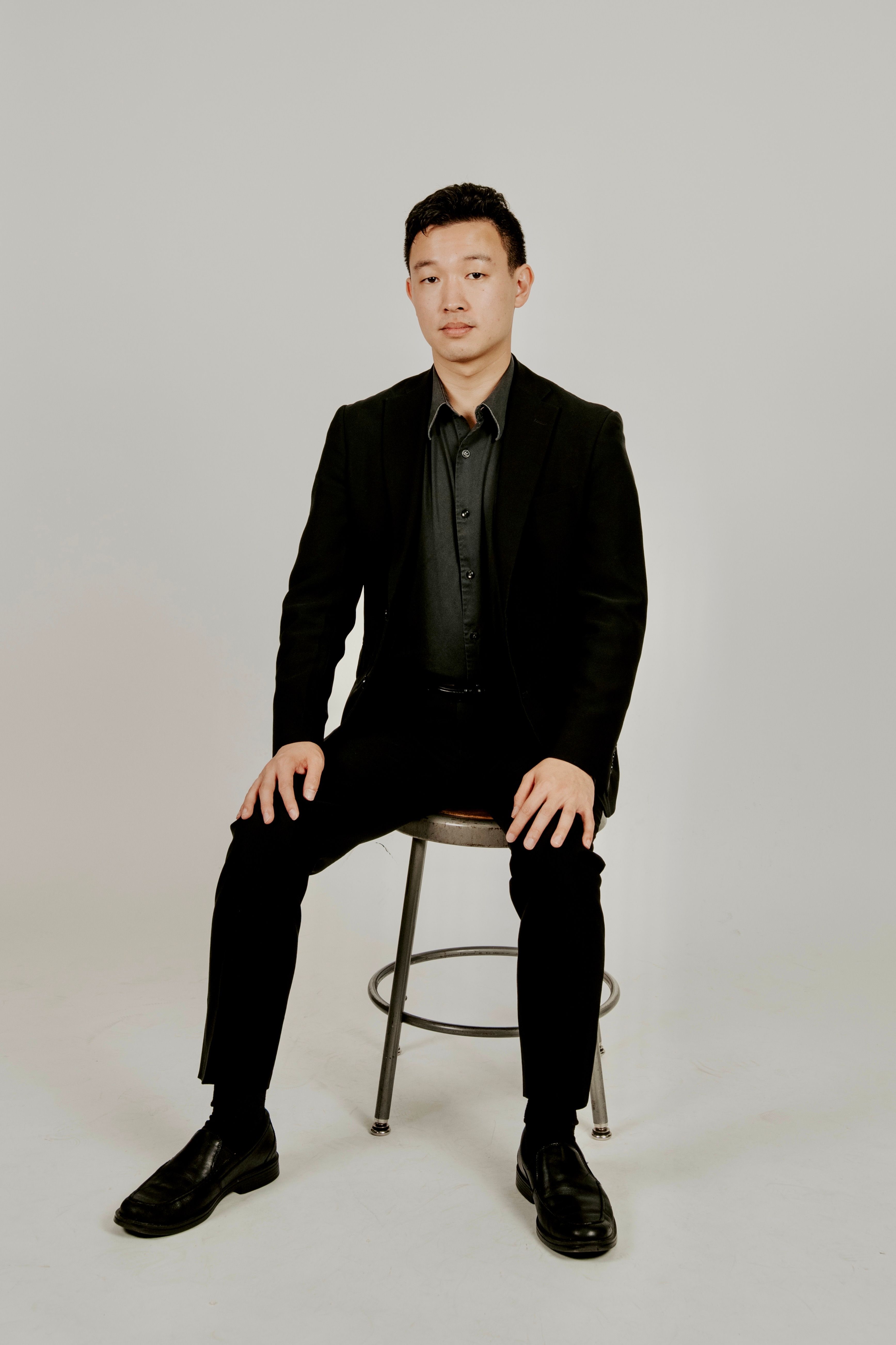 Portrait of a man in a black suit sitting on a stool against a neutral background.