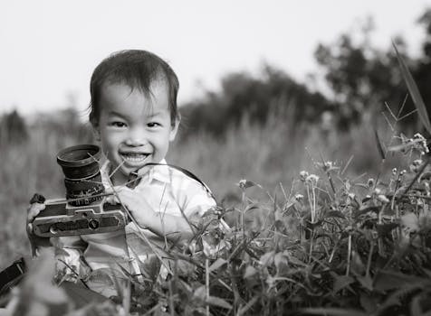 Black and white photo of a smiling child holding a vintage camera in a field of flowers.