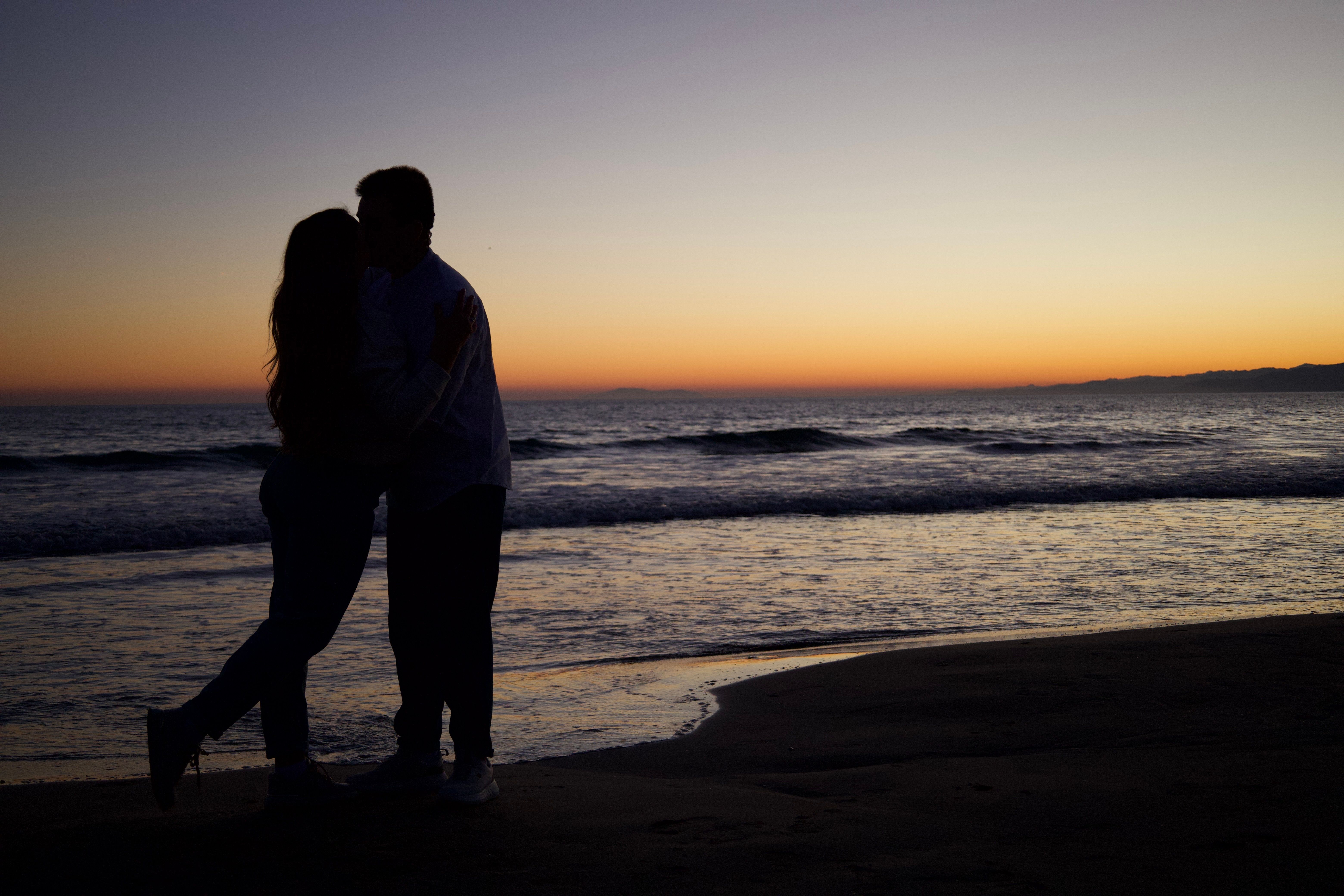 Gratis Silhouette di una coppia abbracciata su una spiaggia al tramonto, immortalando un momento romantico. Foto a disposizione