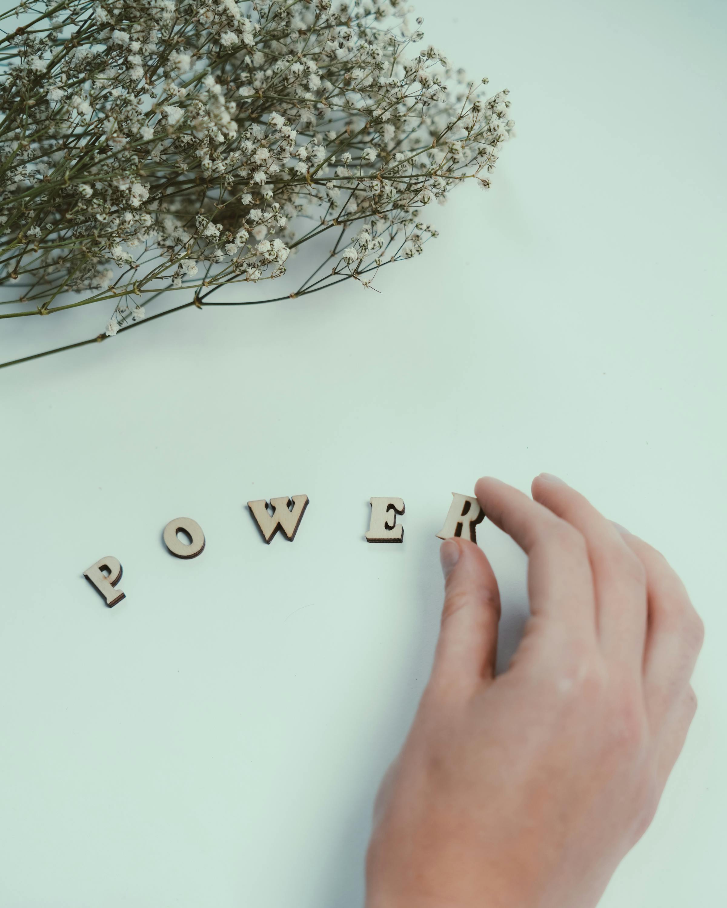 A hand arranging the word 'POWER' with delicate white flowers on a minimalist background.