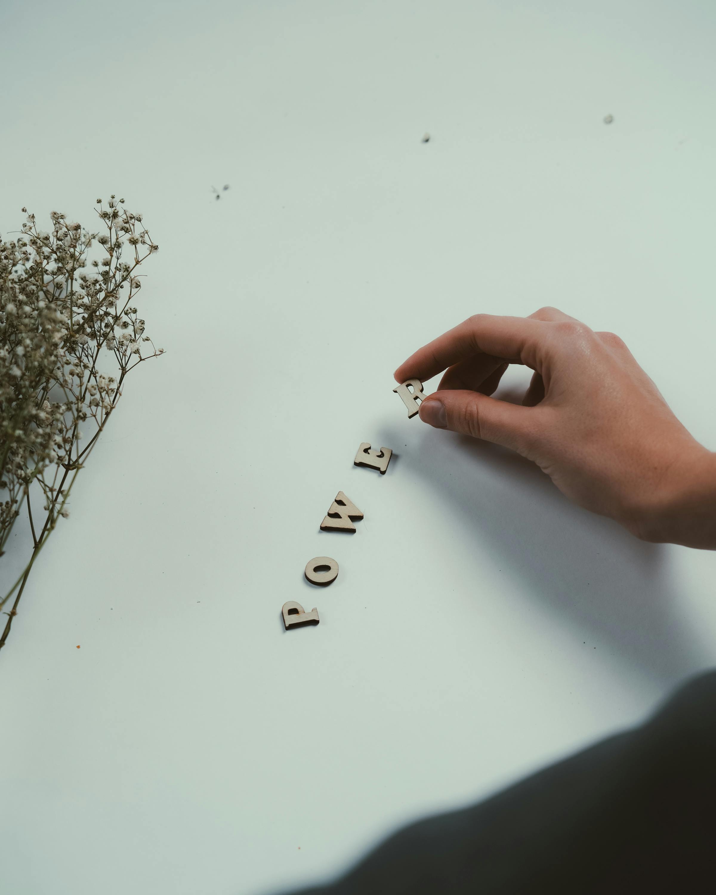 grátis Uma mão organizando letras de madeira formando a palavra "poder" em um fundo suave com flores secas. Foto profissional