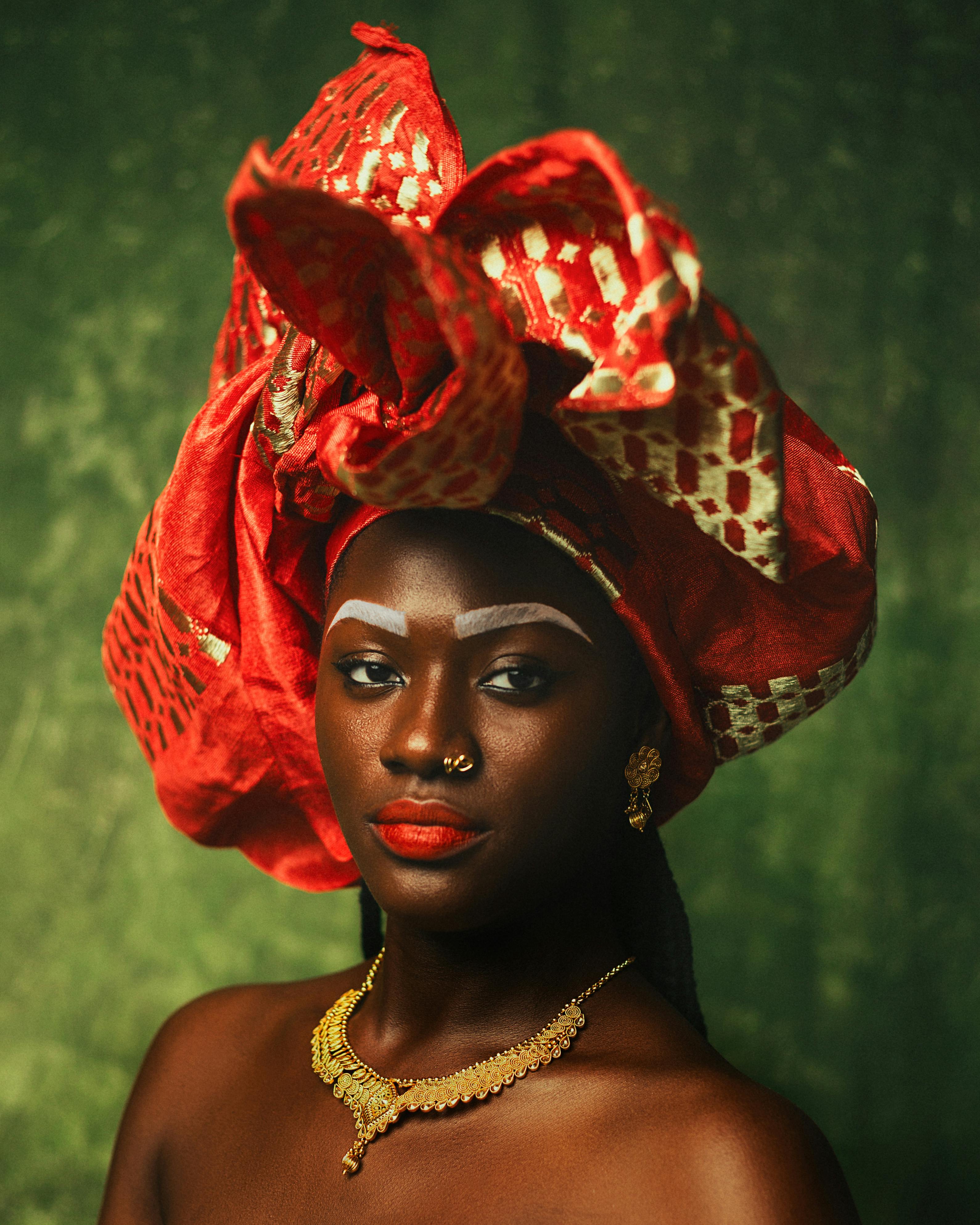Elegant portrait of a woman wearing a vibrant red gele and gold jewelry against a green background.