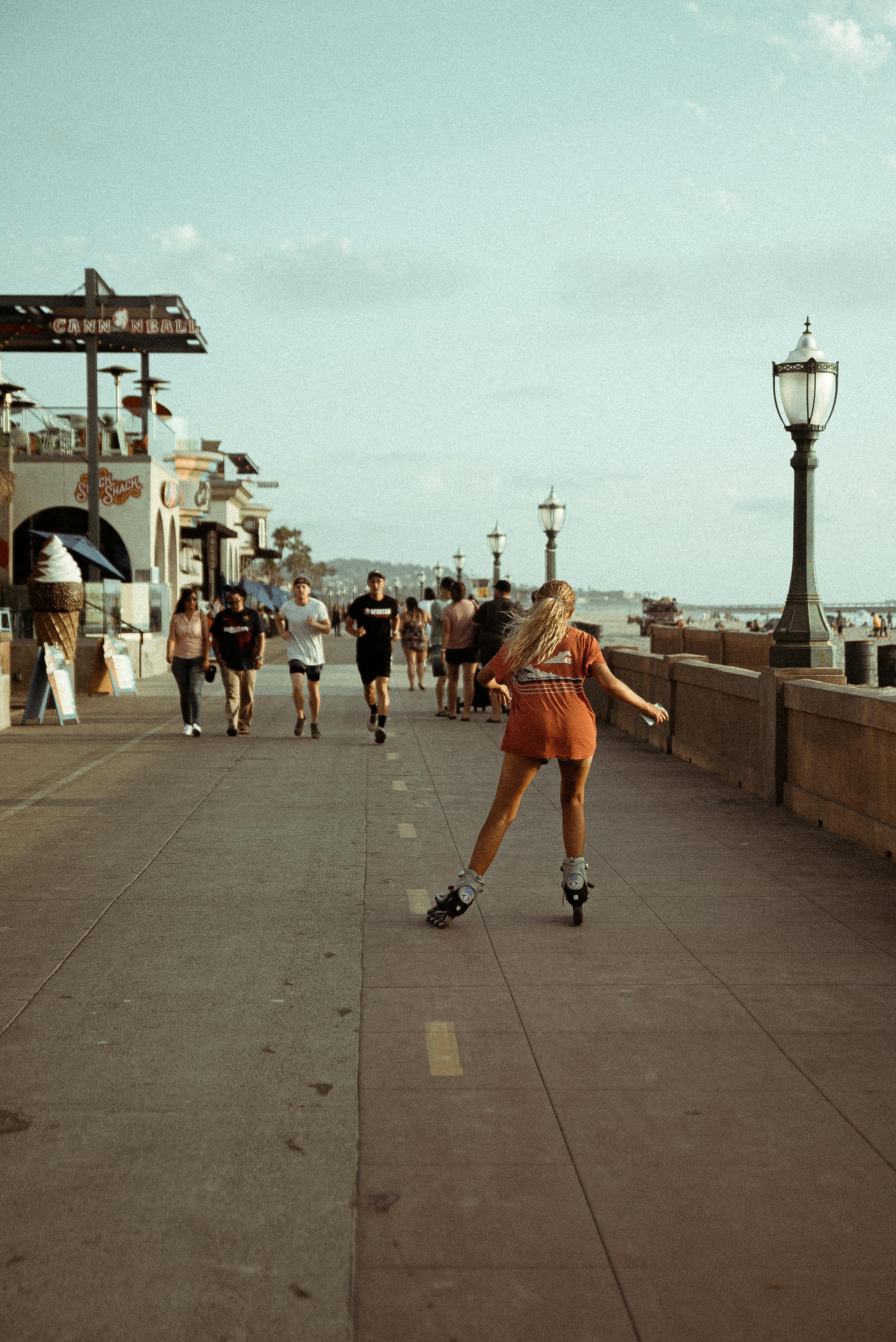 Woman roller skating on a sunny beachside promenade, surrounded by pedestrians and classic architecture.