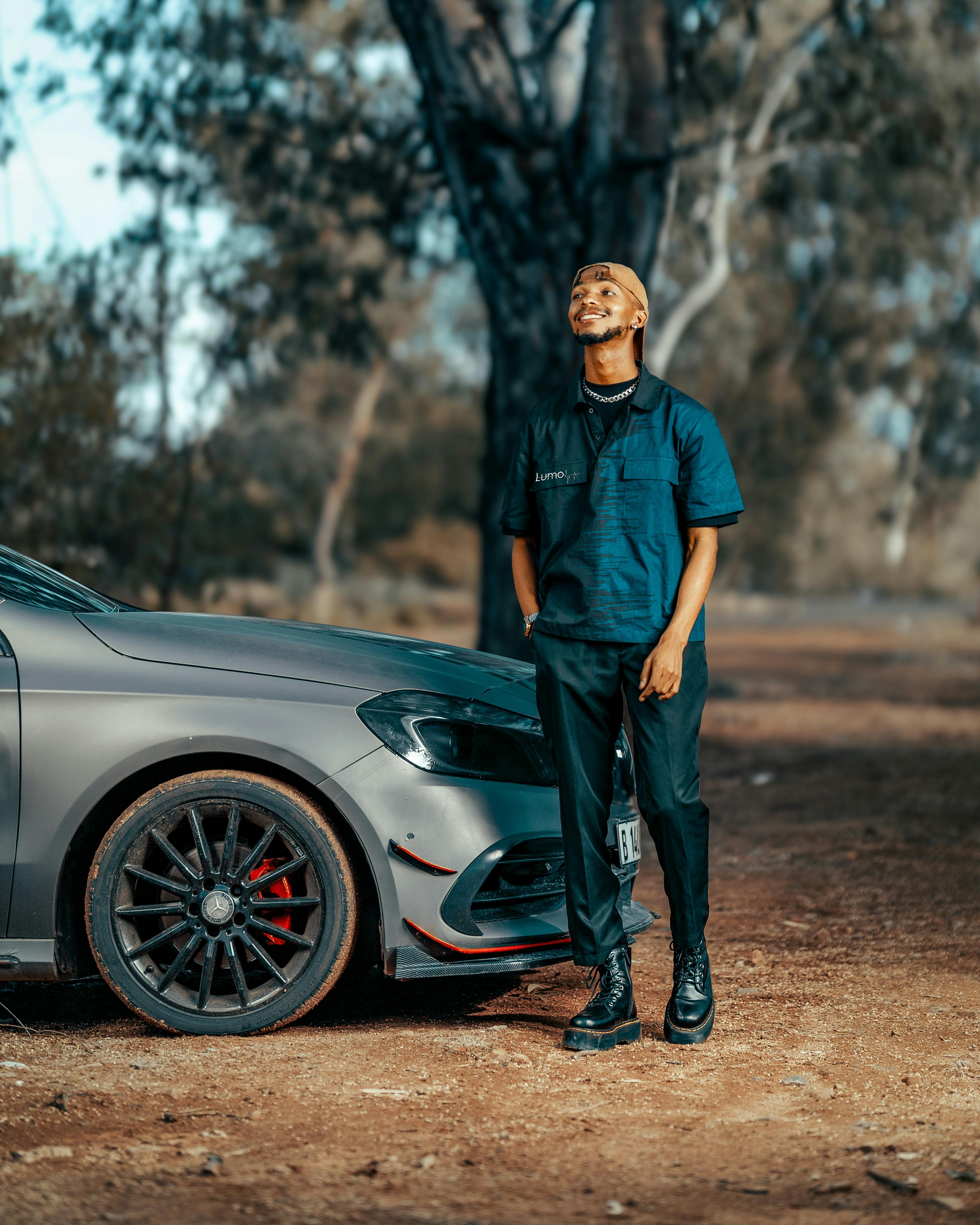 A fashionable man stands confidently beside a sleek luxury car amidst a natural outdoor setting.