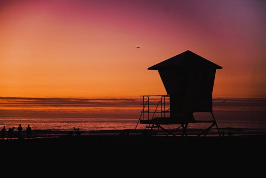 夕阳下，救生员塔的剪影映衬着人们享受海滩宁静的画面，非常适合旅行和休闲主题