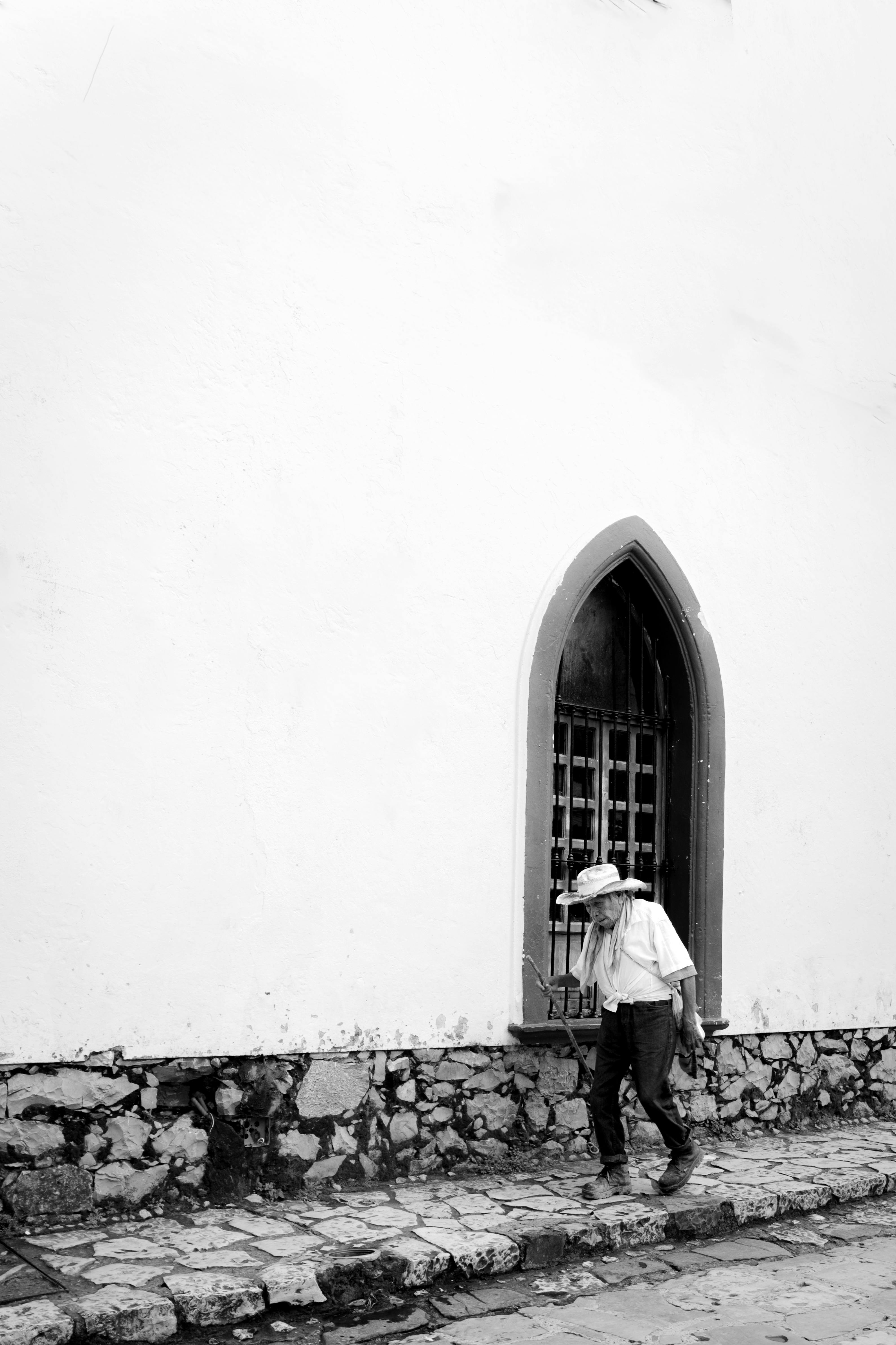 Gratis Fotografía en blanco y negro de un hombre mayor caminando junto a un edificio histórico con una ventana arqueada. Foto de stock