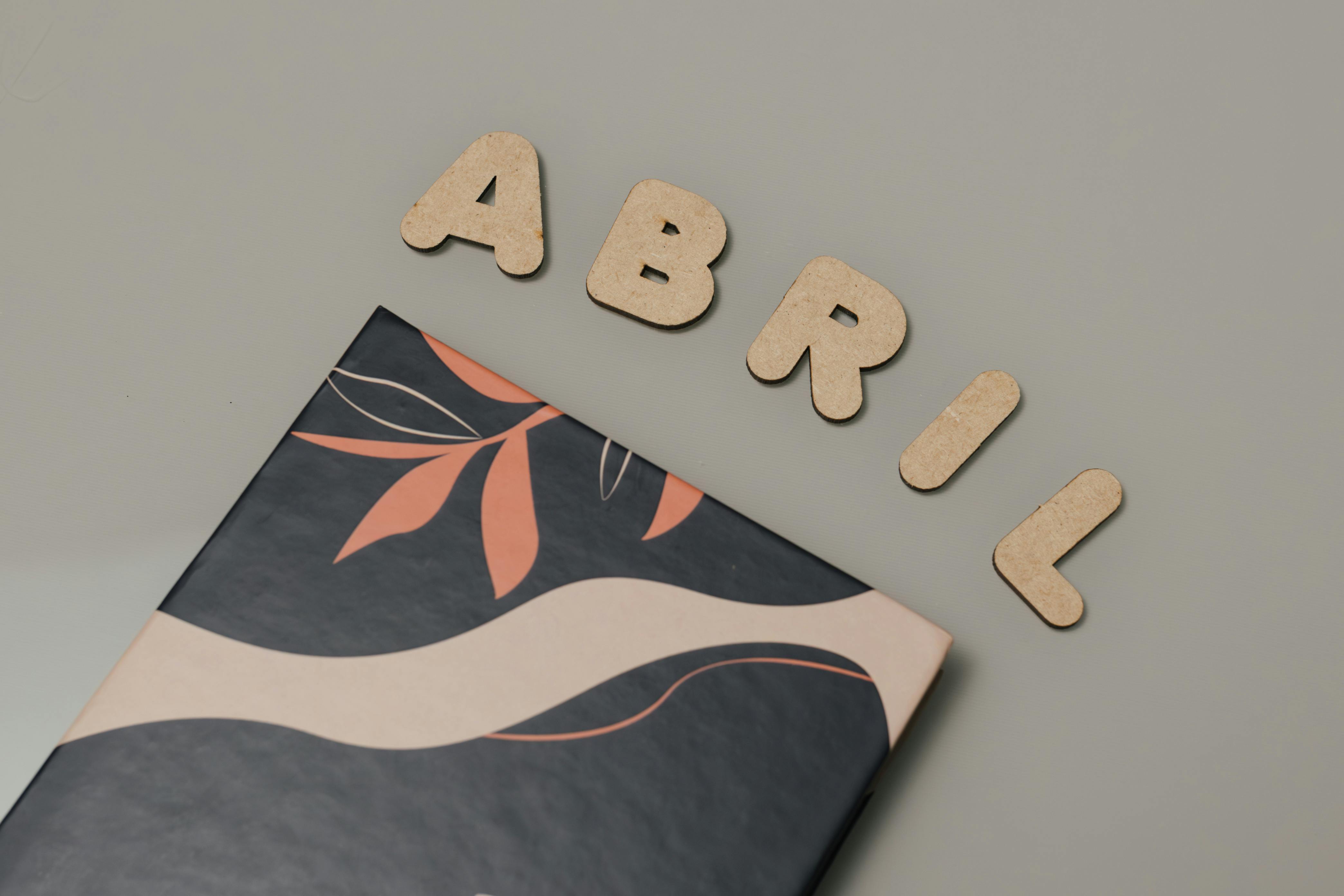 Gratis Huruf kayu yang mengeja Abril pada permukaan datar dengan sampul seni abstrak di bawahnya, menciptakan estetika modern. Foto Stok