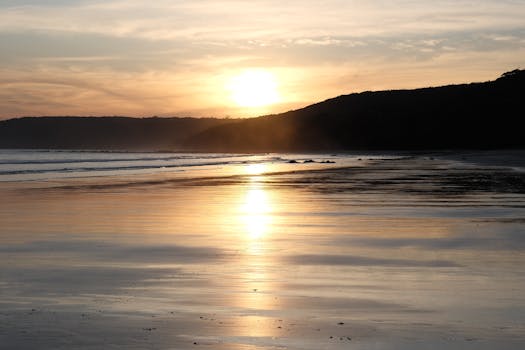 A tranquil beach sunset casting warm reflections over calm waters and silhouetted hills.