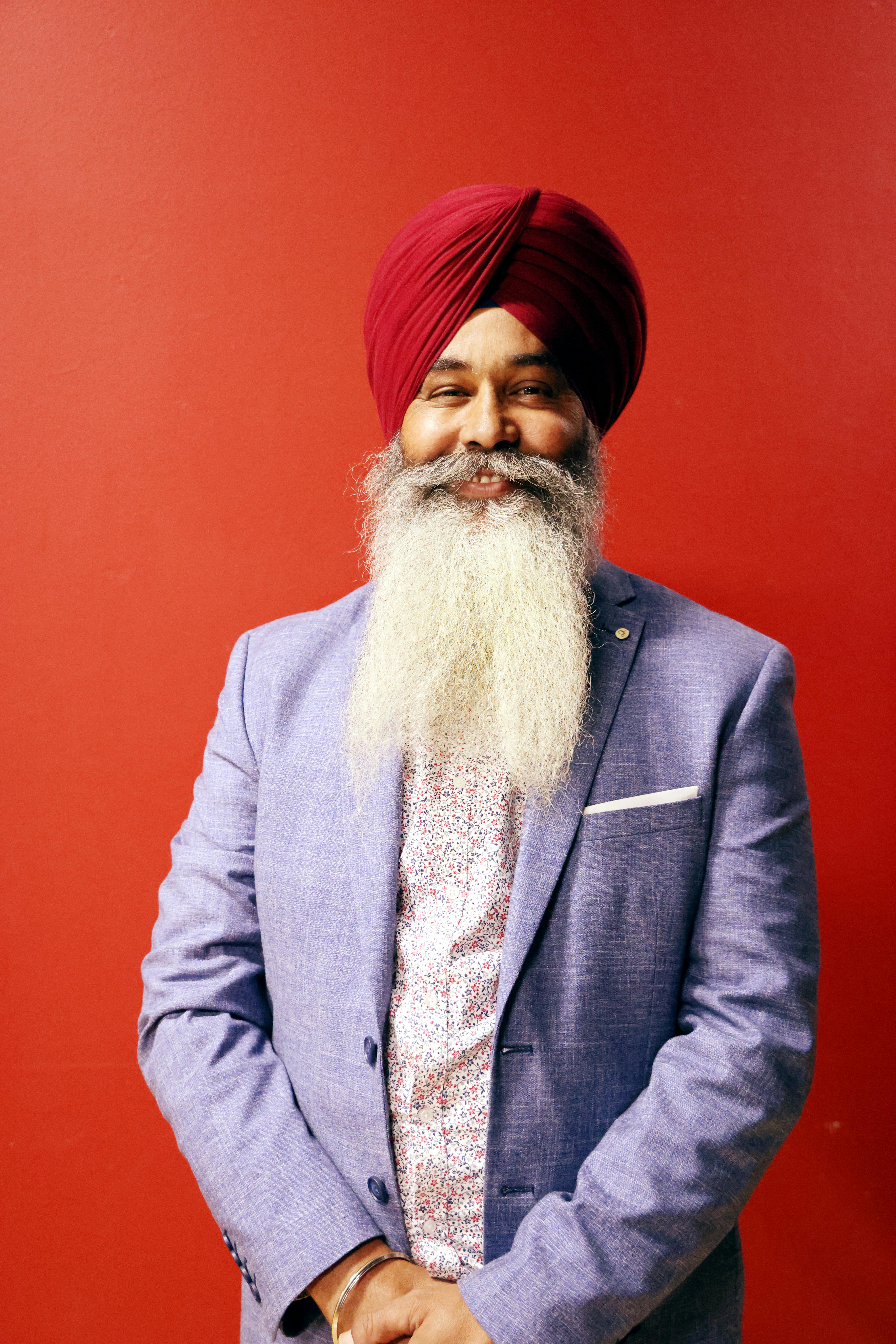 Portrait of a smiling Sikh man in a vibrant turban and suit against a red background.