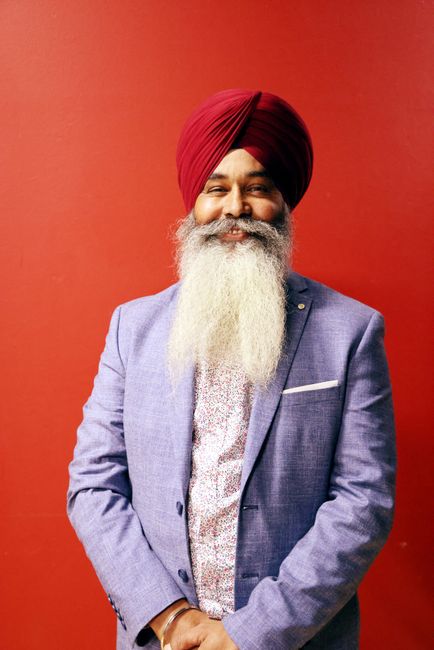 Portrait of a smiling Sikh man in a vibrant turban and suit against a red background.