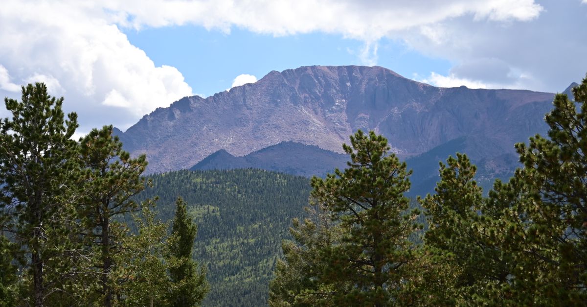 Breathtaking view of Pikes Peak behind a lush green pine forest under a blue sky with clouds.